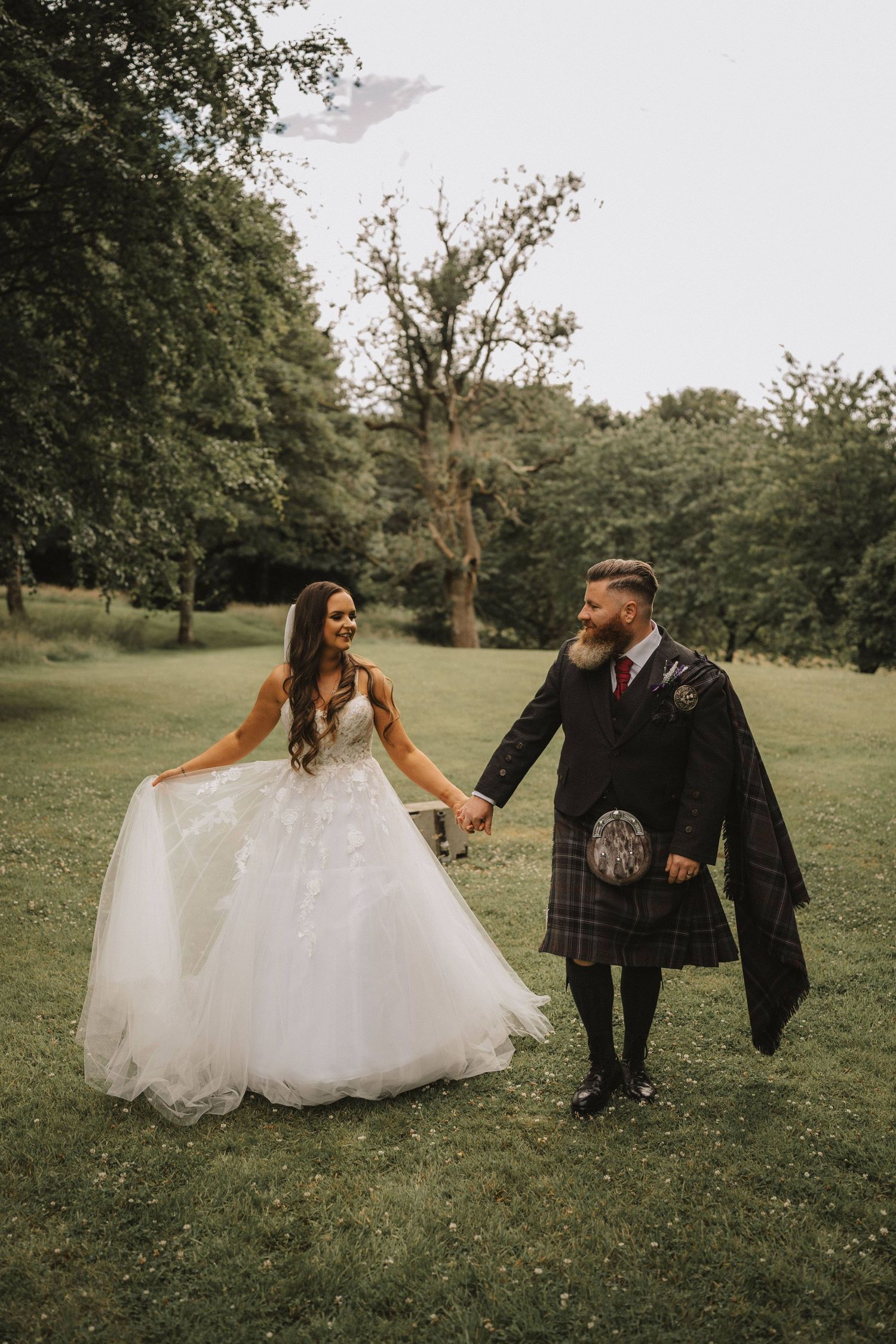 Newlyweds share playful moments on a grassy lawn, with groom in traditional Scottish attire.