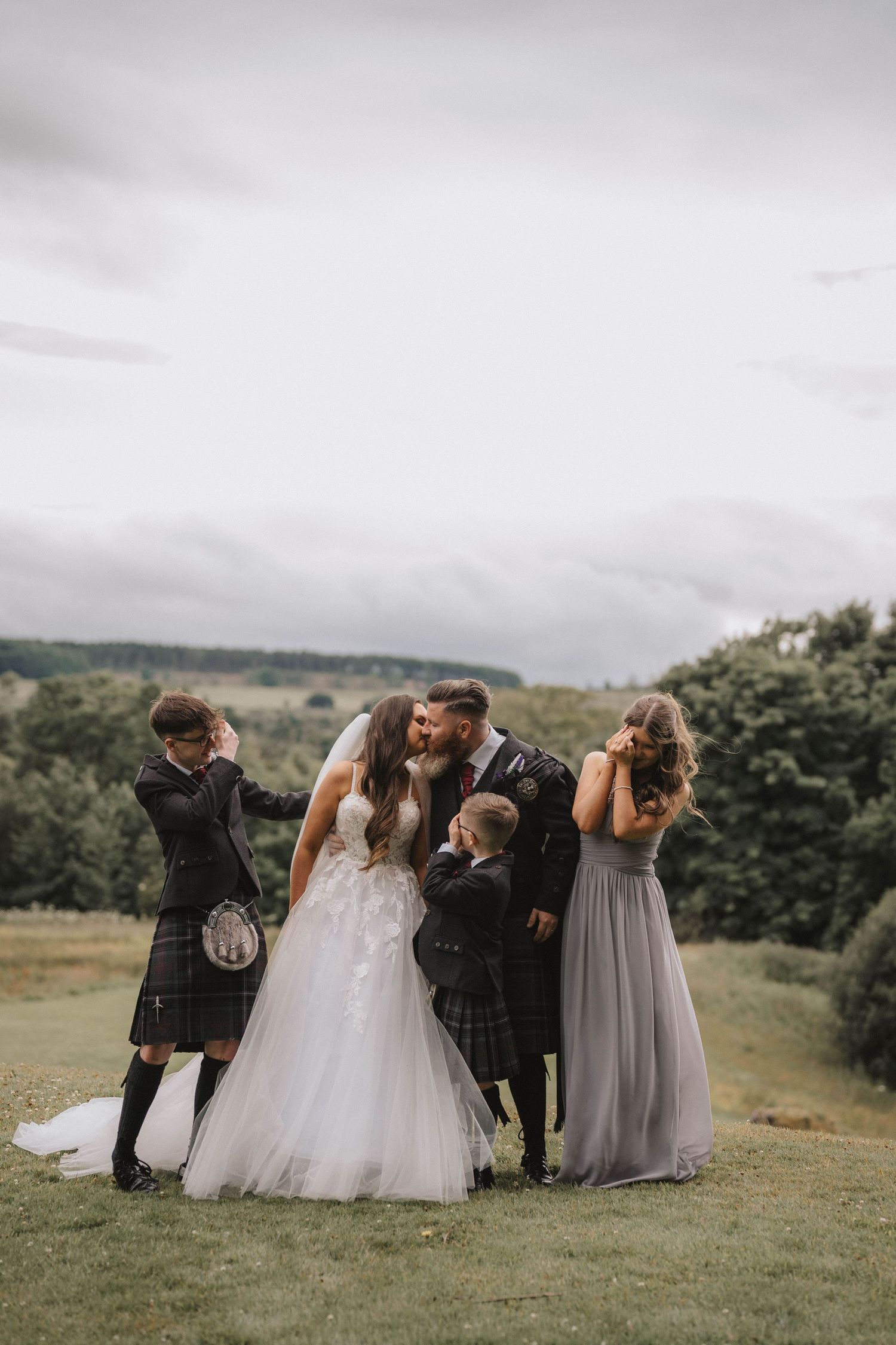 Wedding party lounges on grassy hillside during outdoor photo session.