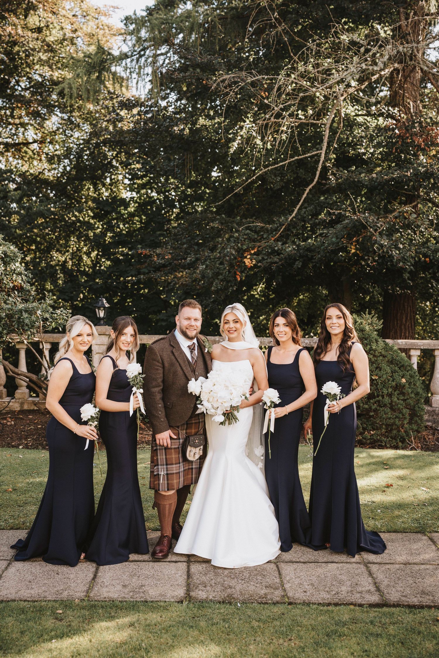 Wedding party in black bridesmaid dresses and brown suit posing together on grassy lawn near trees.