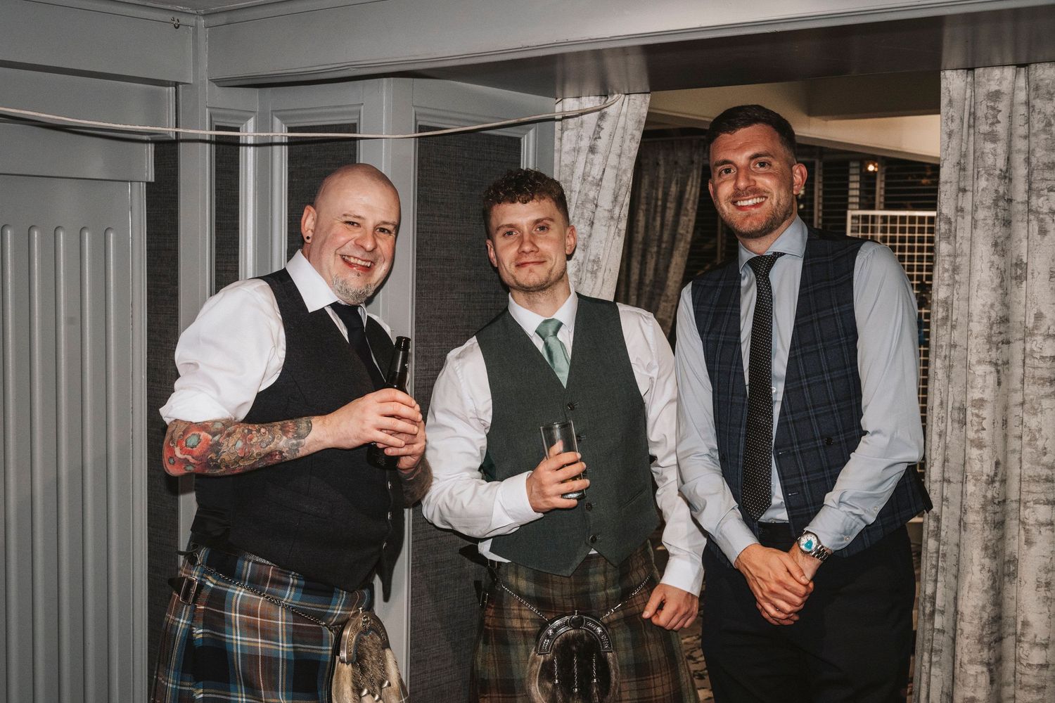 Three men in formal attire including kilts and vests pose together at an indoor event.