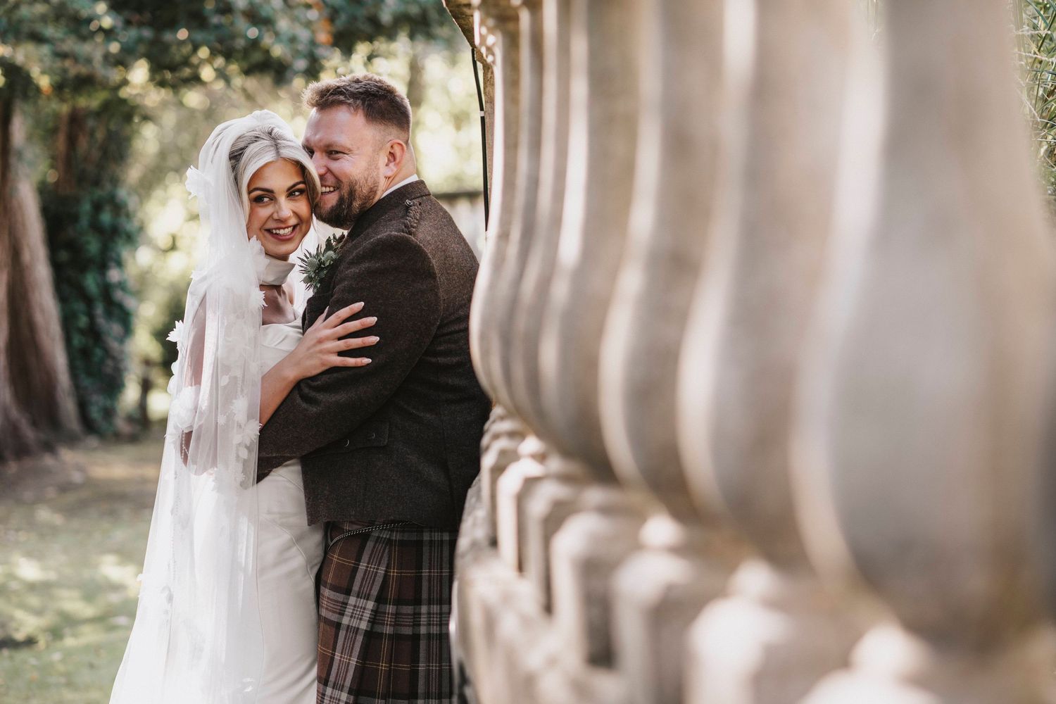 A romantic wedding couple embraces outdoors while surrounded by elegant stone columns and natural light.