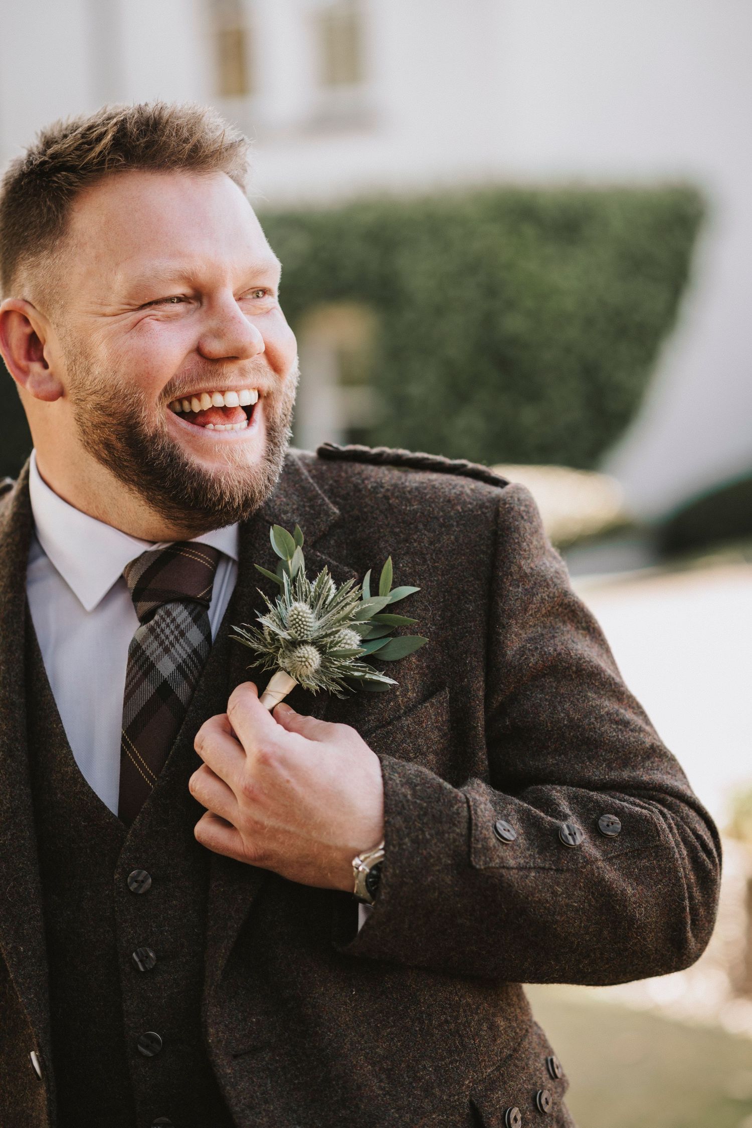 A person wearing a brown tweed suit jacket smiles while adjusting their boutonniere in a series of candid photos.
