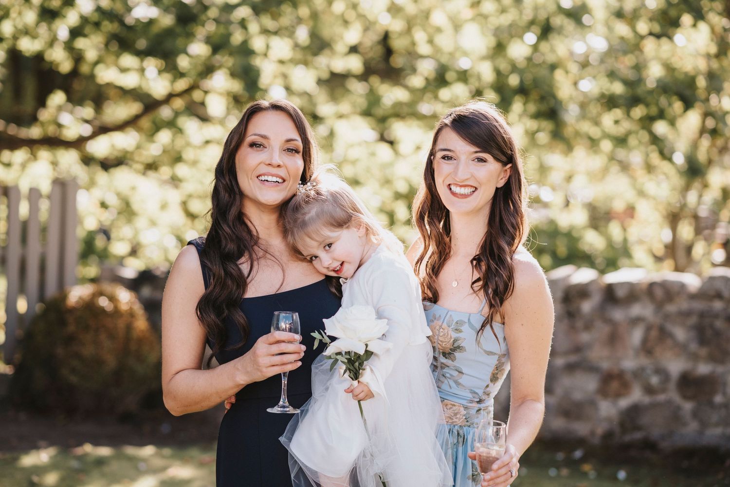 Wedding party members laugh and smile together while holding bouquets in an outdoor garden setting.