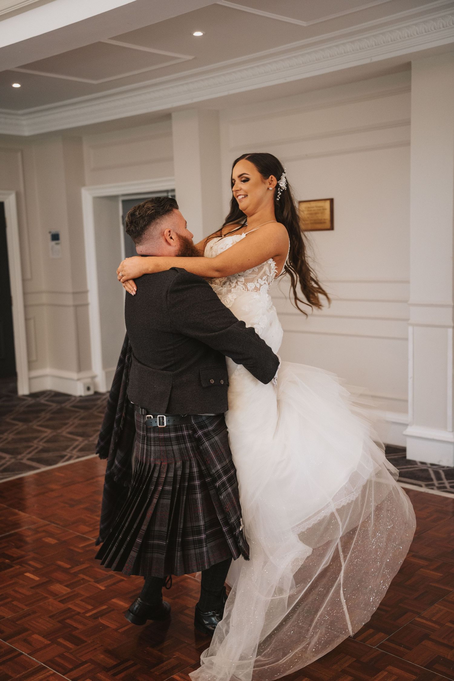 A couple shares their first dance at a wedding reception, with the groom wearing a traditional Scottish kilt.