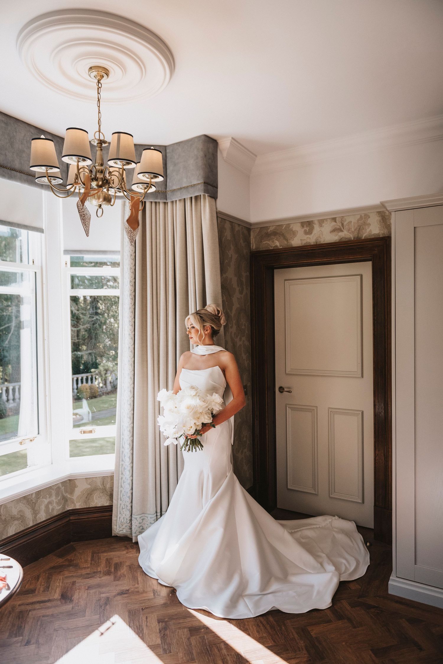 A bride in a flowing white gown twirls in a room with herringbone wood floors and elegant cream curtains.
