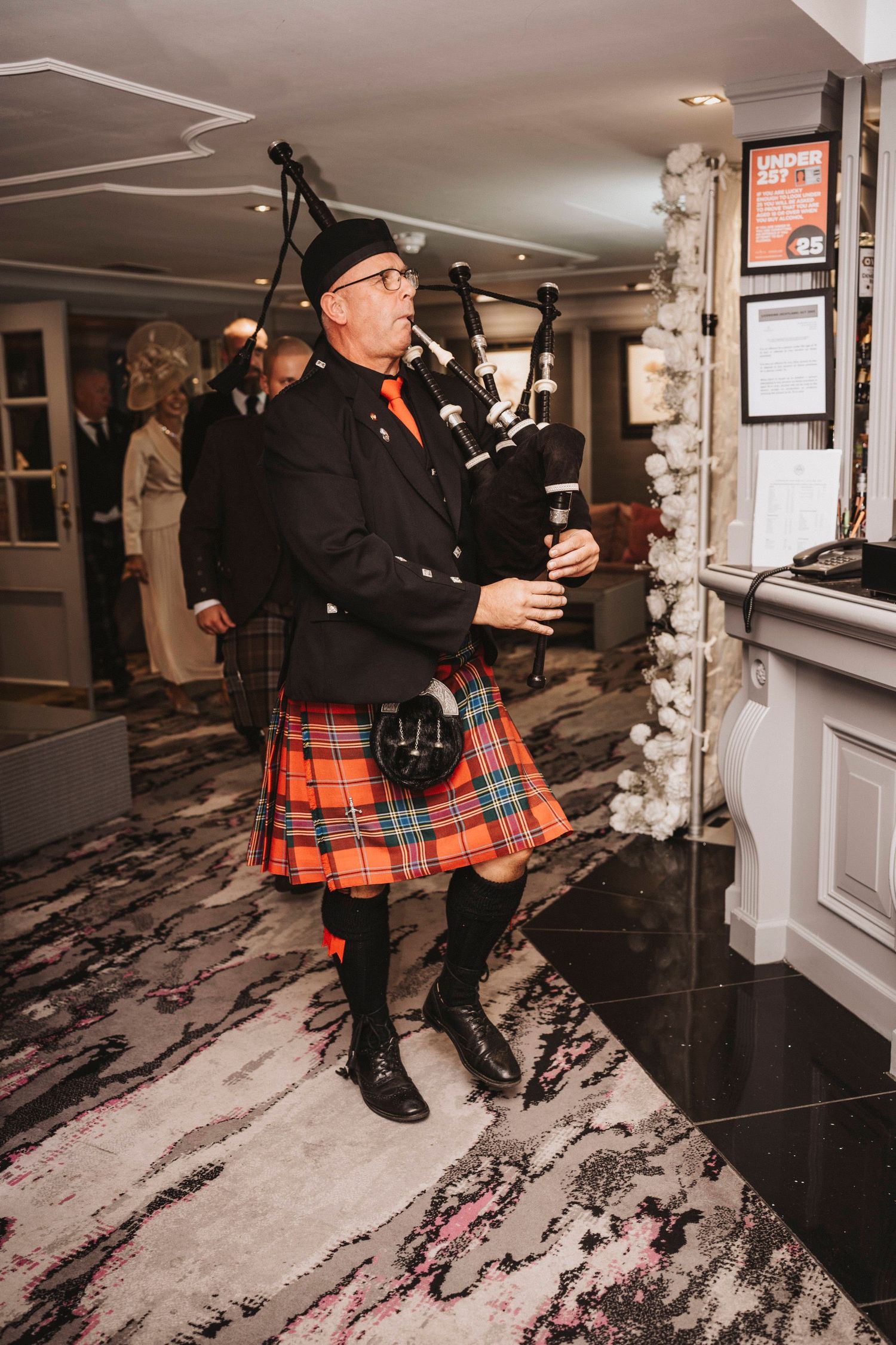 A bagpiper in traditional Scottish attire performs in a formal indoor venue with marble floors and decorative lighting.