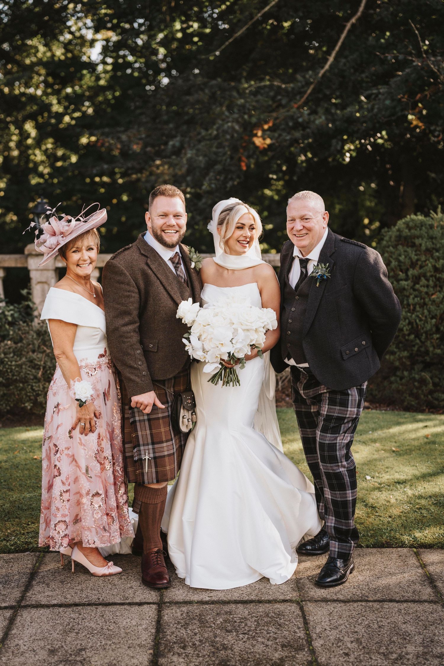 A traditional Scottish wedding party poses outdoors with the bride in white and men wearing kilts and formal attire.