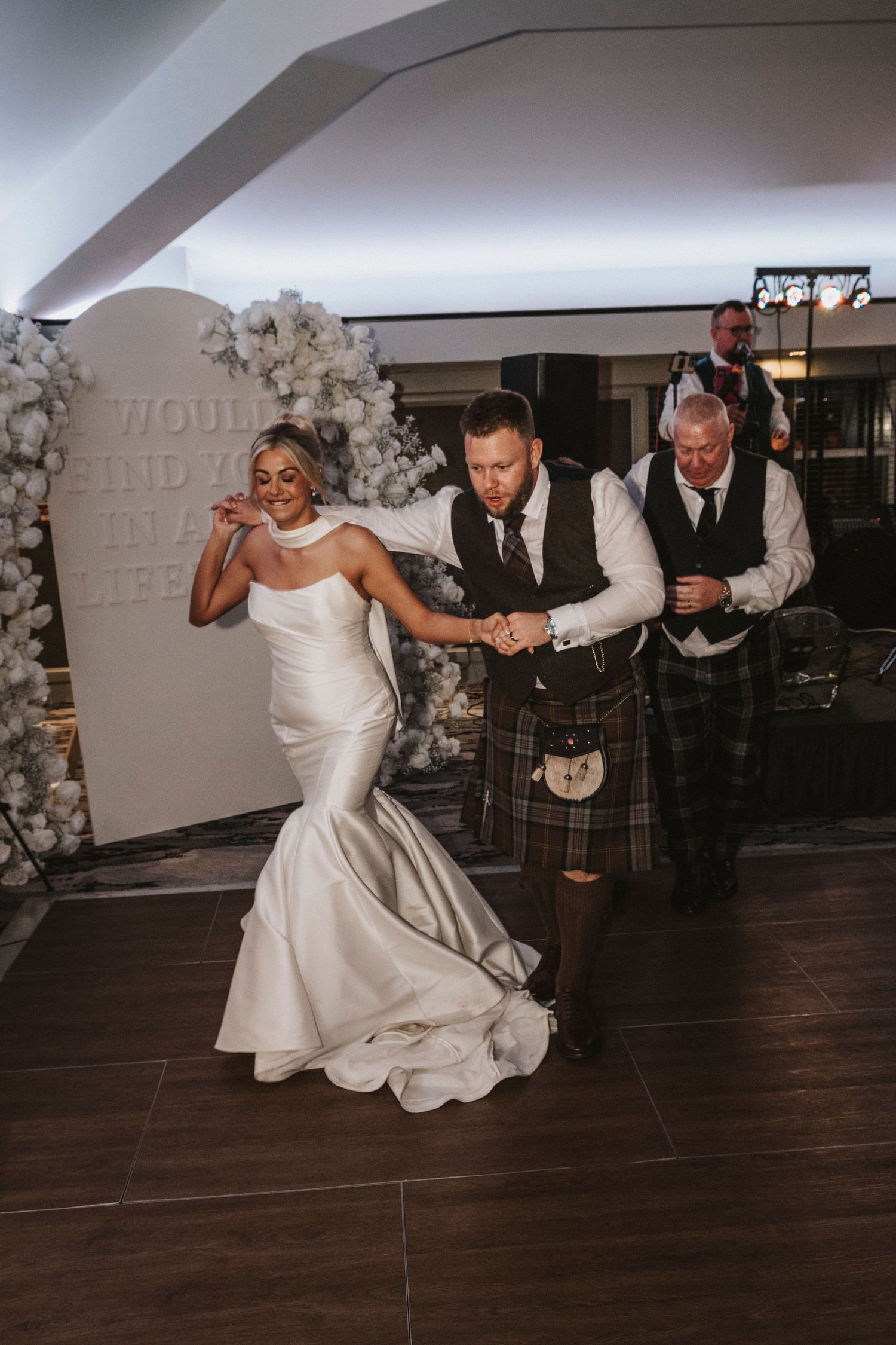 A couple performs an energetic dance sequence on a dark wooden floor at their wedding reception.