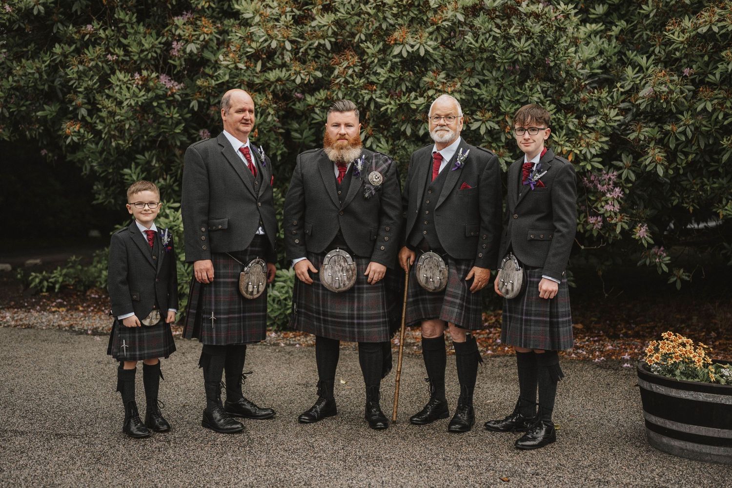 Wedding party members in dark suits pose together on brick steps near autumn foliage.