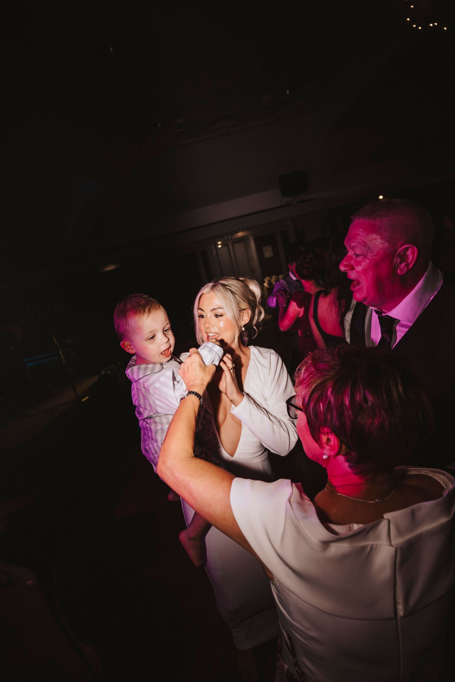 Wedding reception dance floor scene with dramatic pink lighting and people dancing in low light conditions.