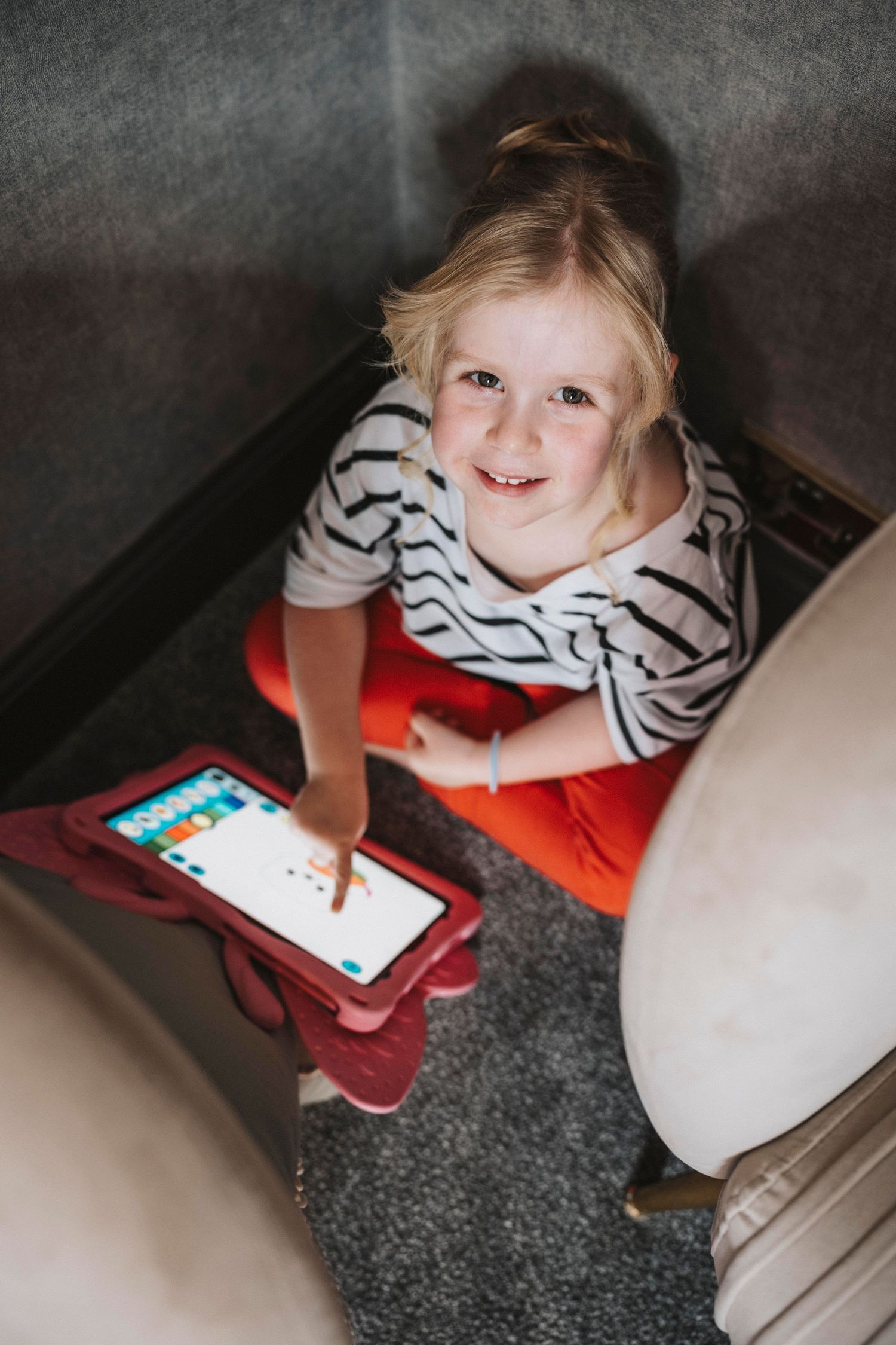 A child in a striped shirt and red pants uses a tablet device while sitting on gray carpet.