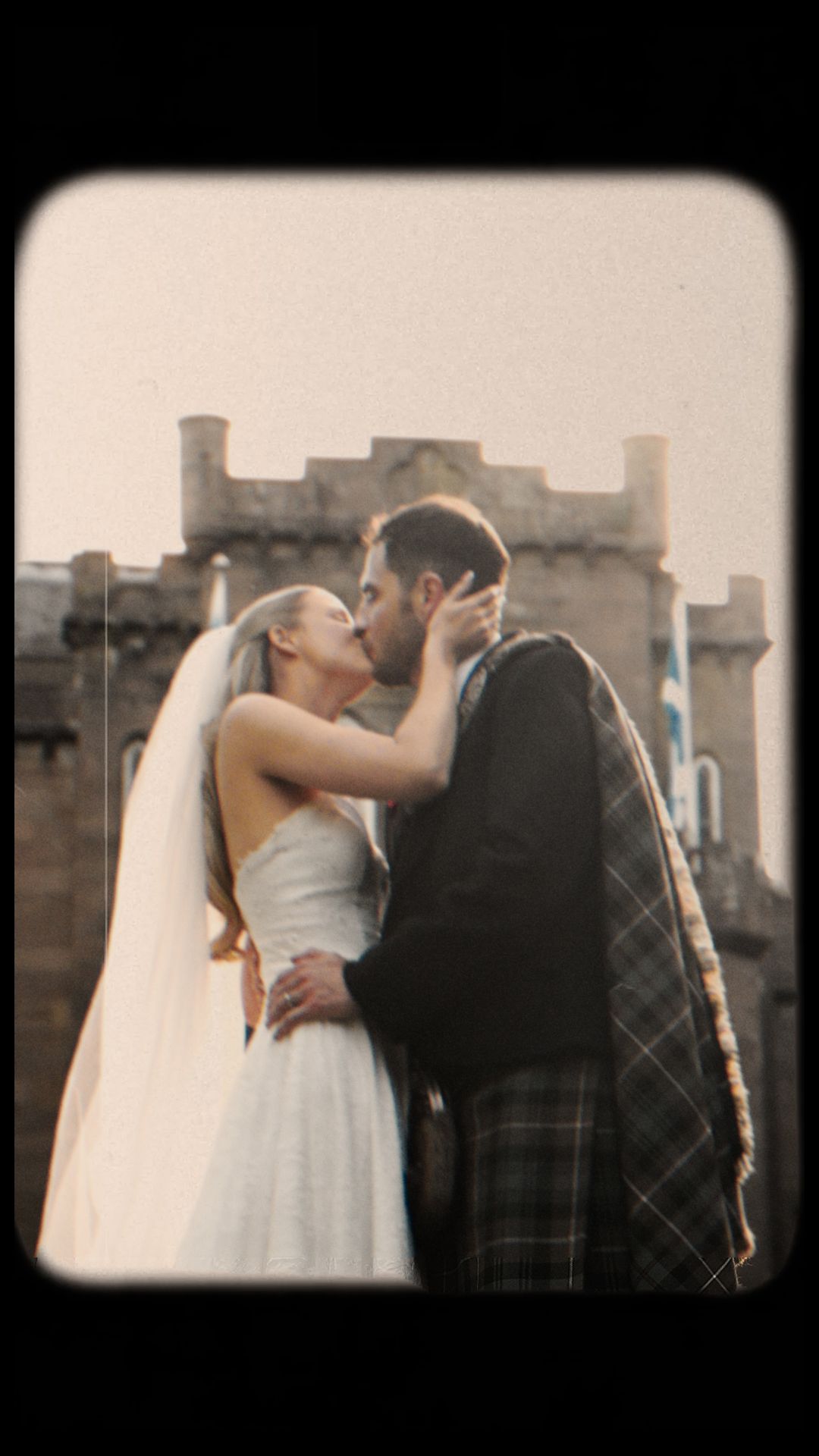 A bride and groom share a romantic kiss in front of a Scottish castle, the groom wearing a traditional tartan kilt.