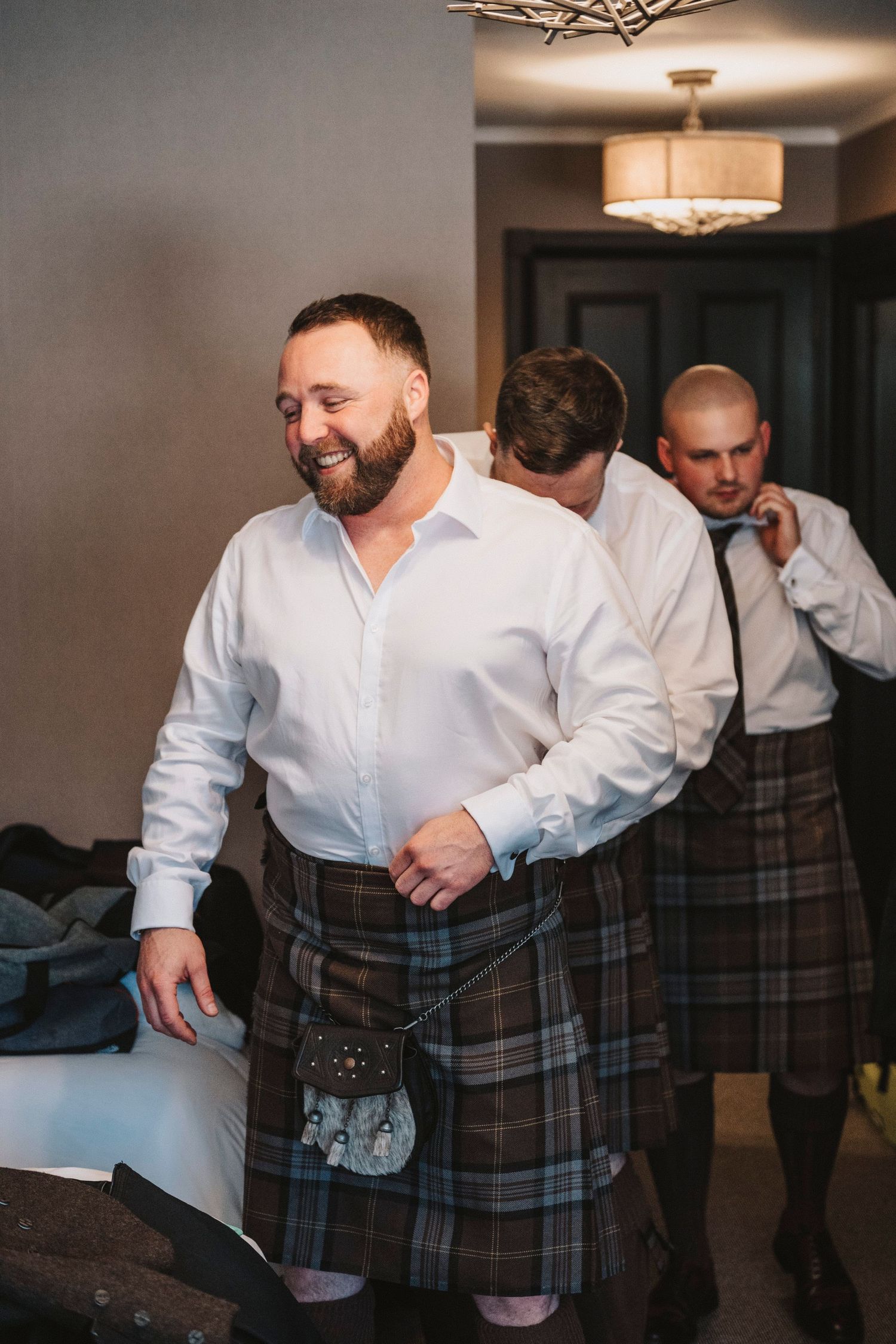 People wearing traditional Scottish kilts and white dress shirts prepare for a special occasion indoors.