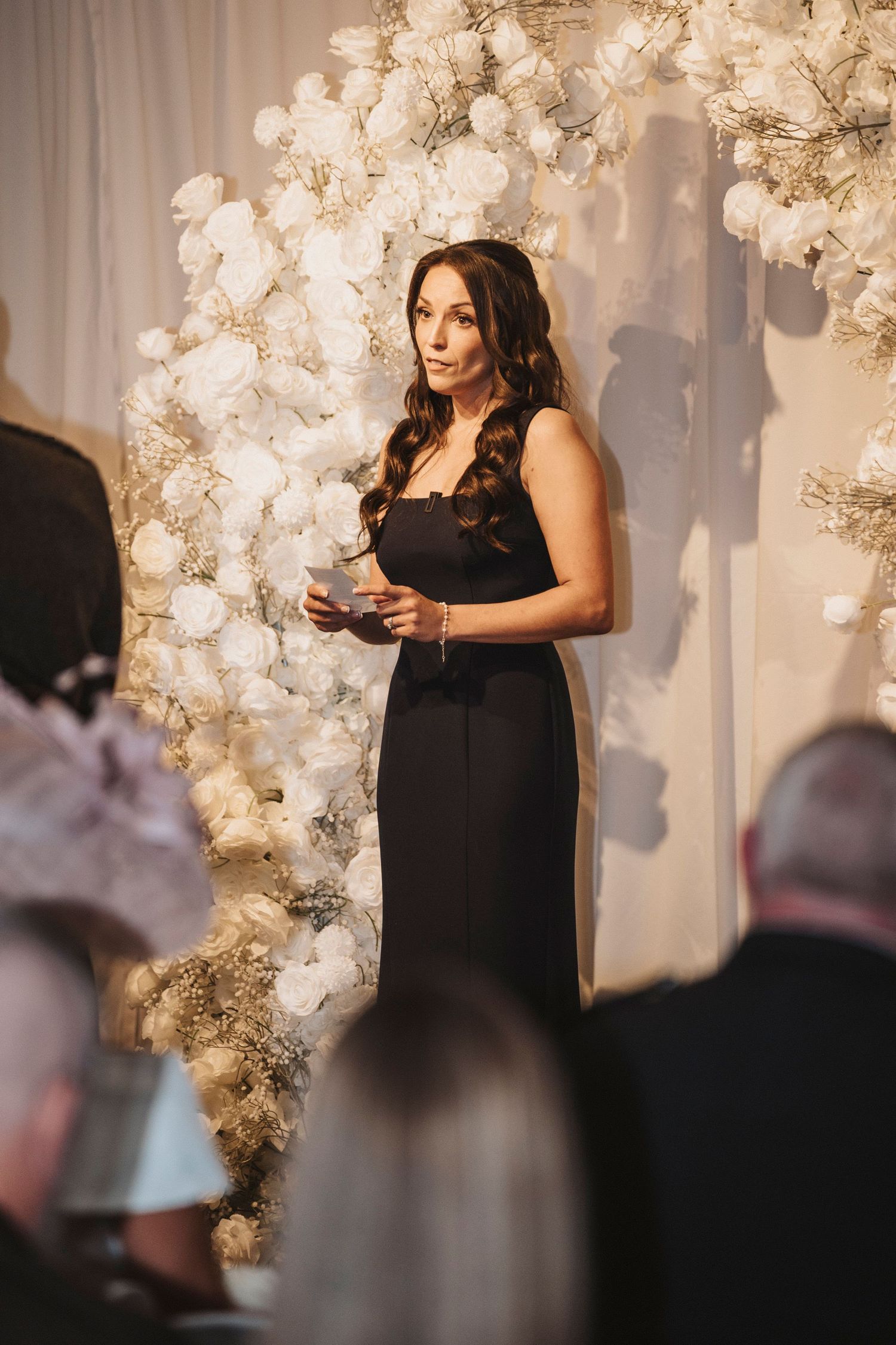 A person in a black formal dress speaks at a podium with elegant white floral decorations in the background.