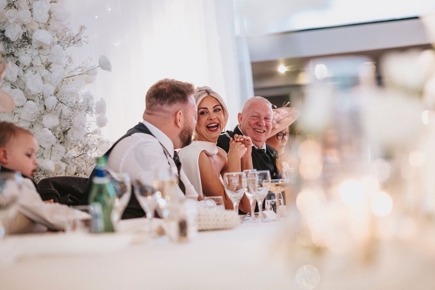 Wedding guests seated at elegant tables with white floral centerpieces share joyful moments during a reception celebration.