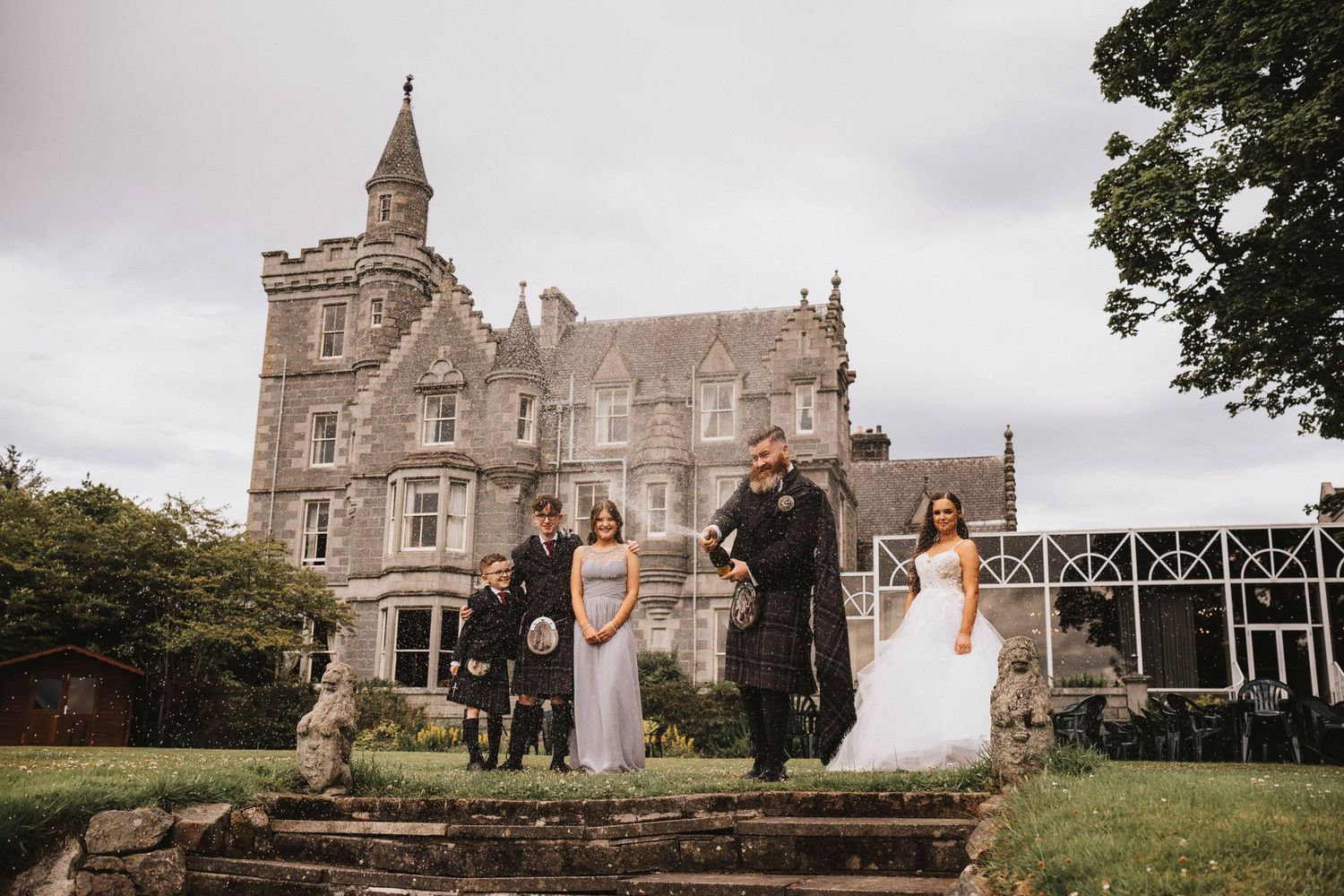 A wedding party poses on the steps of a grand Victorian castle estate under an overcast sky.