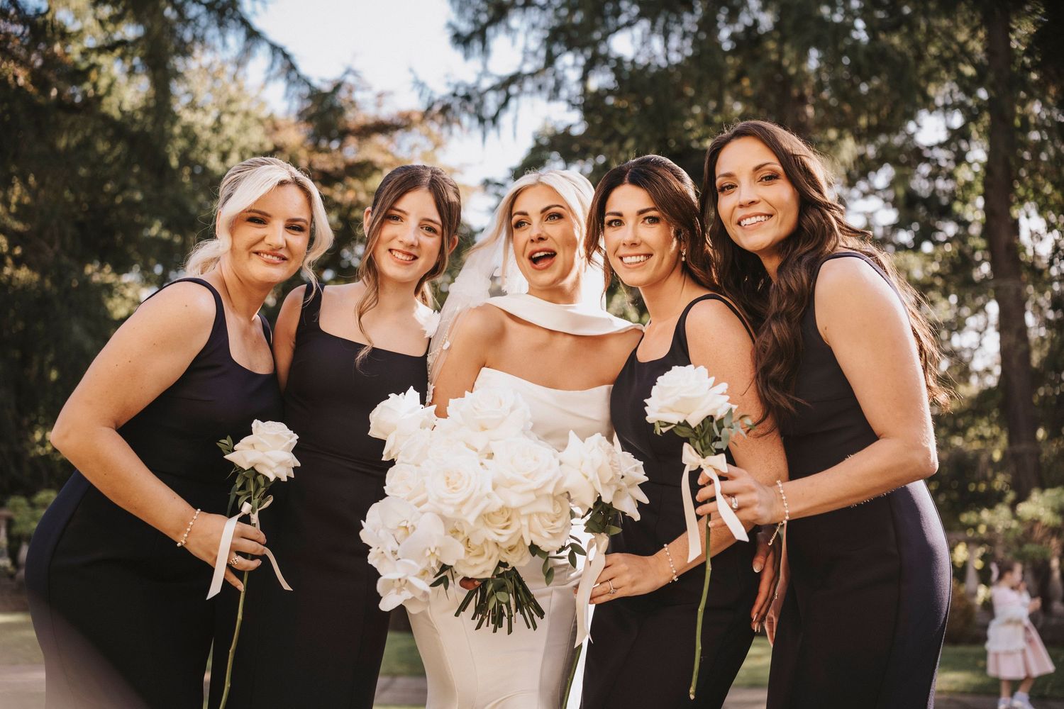 Wedding party in black dresses poses together while holding white floral bouquets during outdoor celebration.