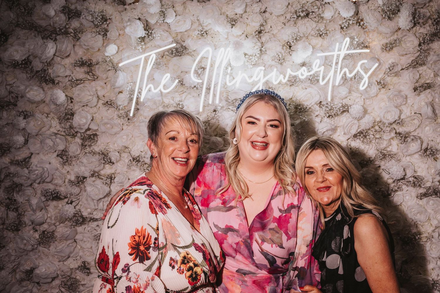 Three people in floral outfits pose together in front of a textured wall at a photo booth event.