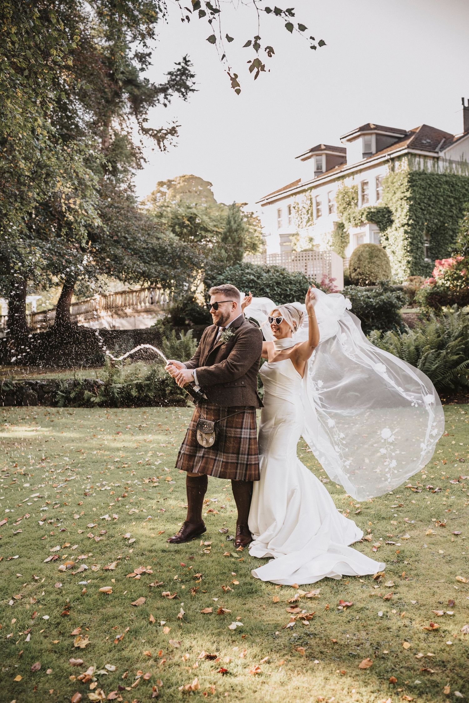 A romantic wedding couple dances and twirls on an autumn lawn with falling leaves and white buildings in the background.