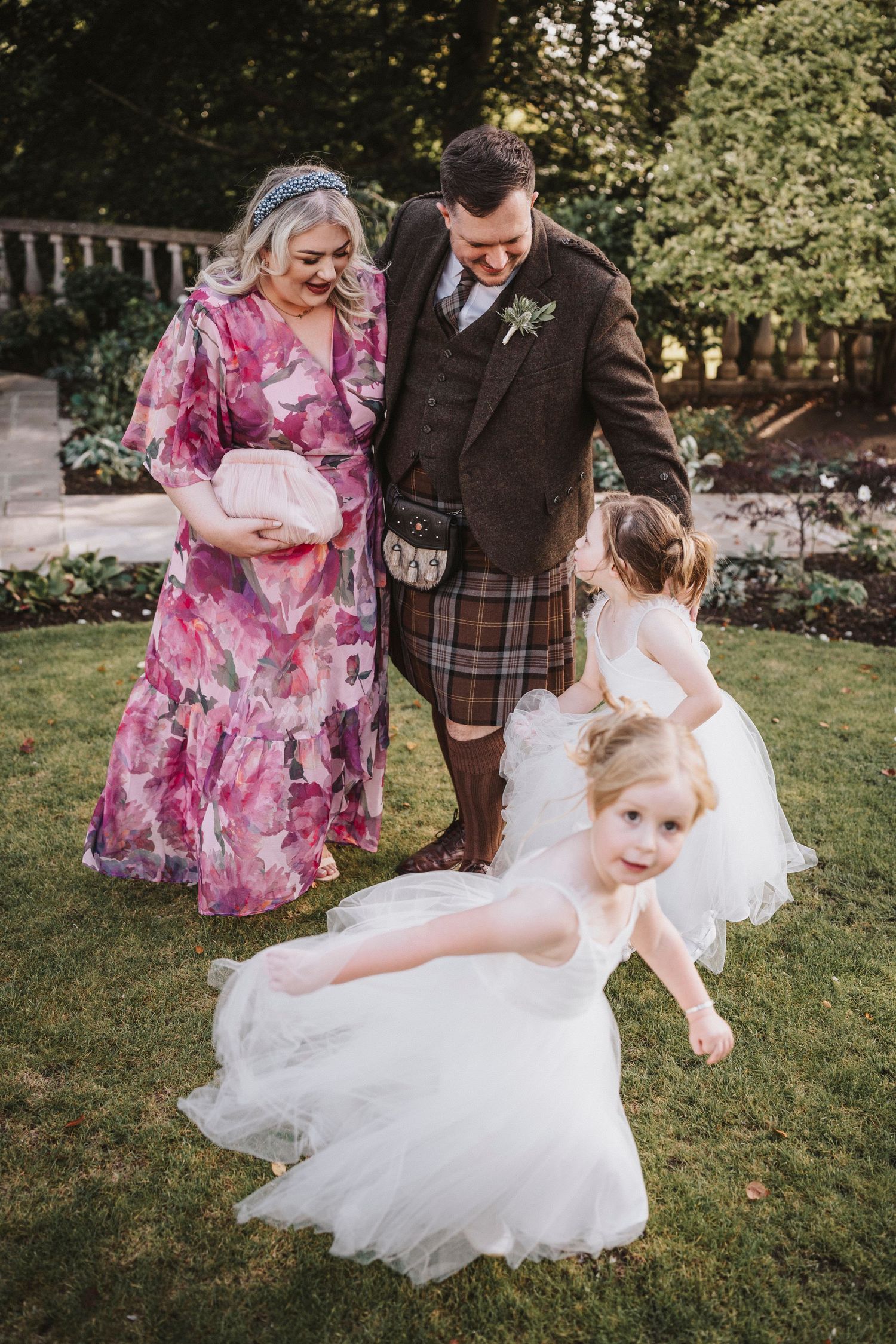 A couple in wedding attire dances and twirls on a sunny lawn surrounded by lush greenery and gardens.