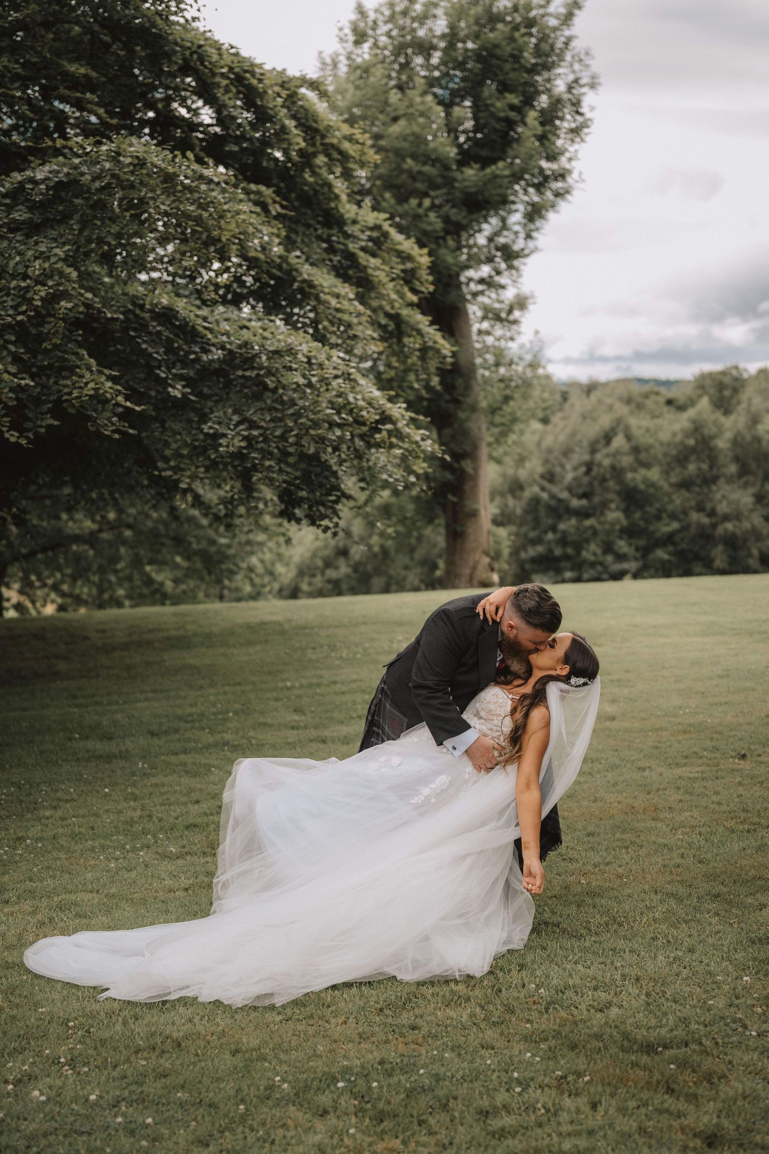 Romantic wedding couple sharing an intimate dip pose on a green lawn surrounded by trees.