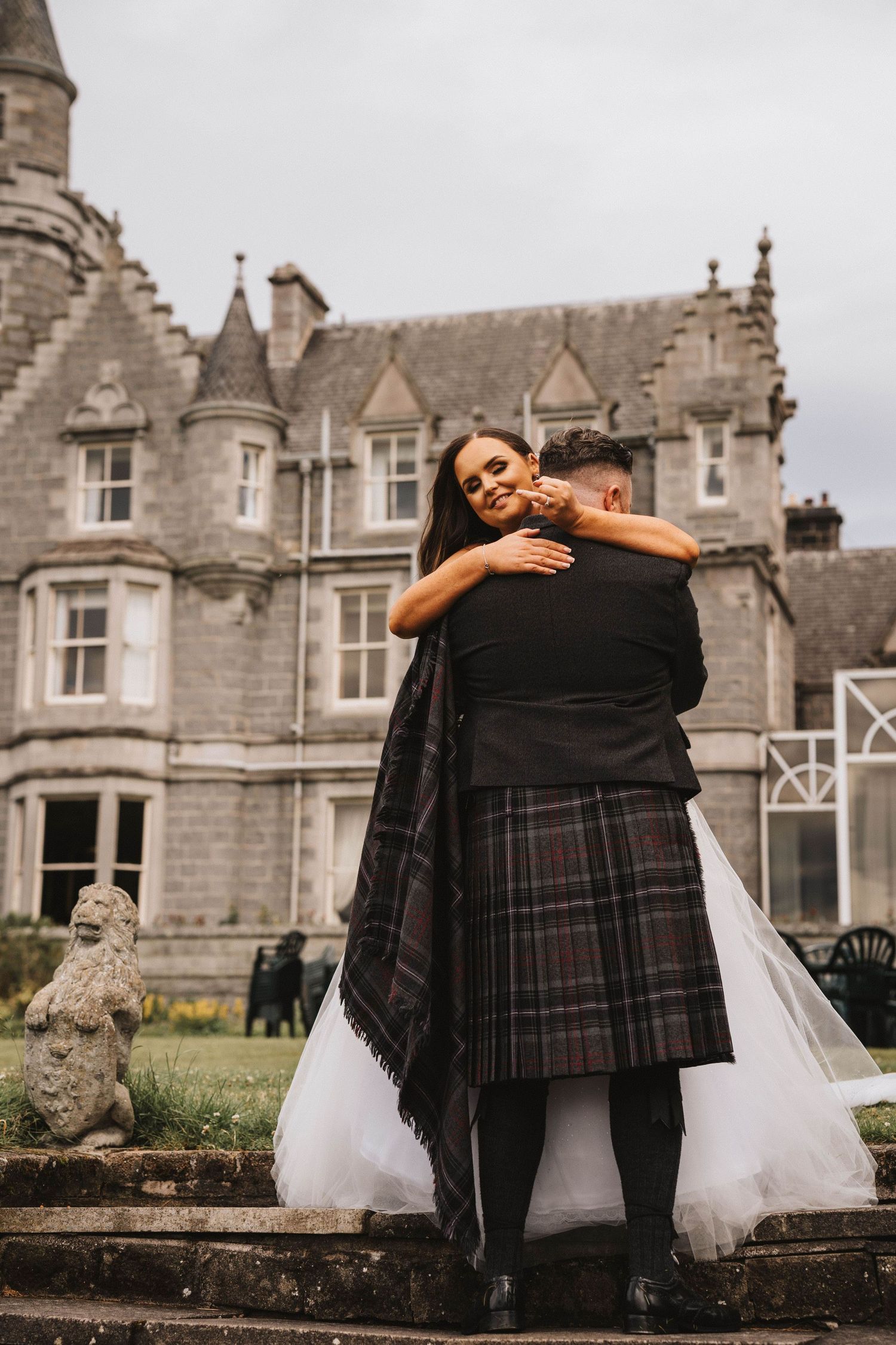 Bride in flowing white gown embraces groom in traditional Scottish kilt outside stone manor house.