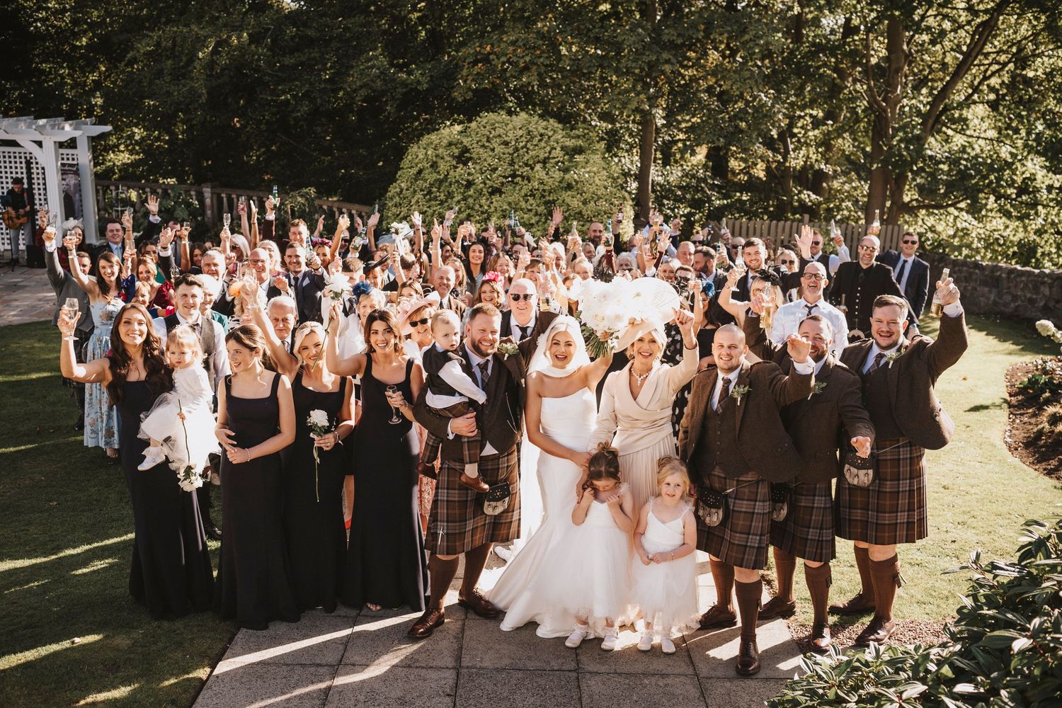 Large wedding party gathered outdoors with guests wearing black attire and traditional Scottish kilts in sunny garden setting.