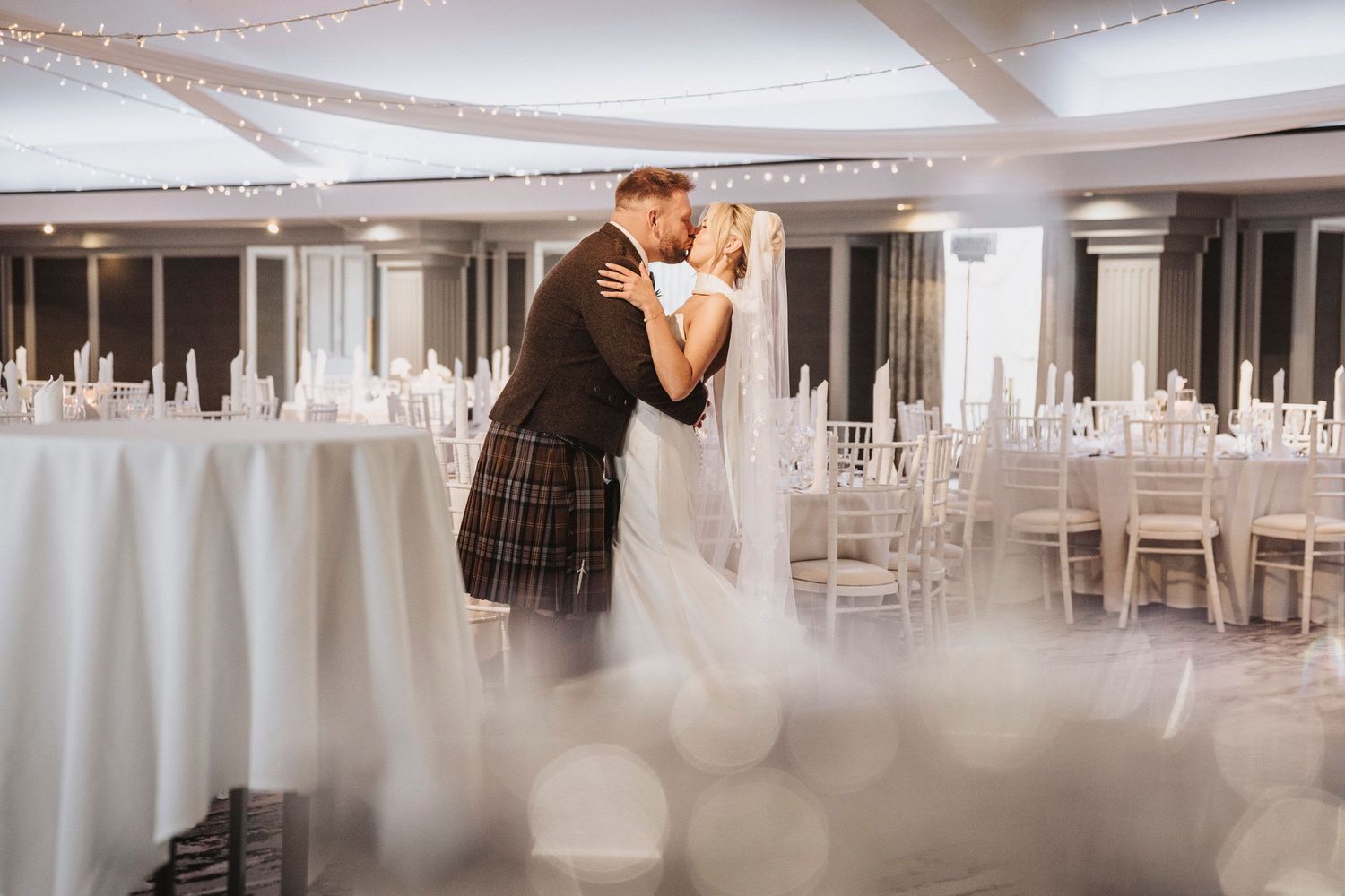 A romantic wedding couple dancing in an elegant white ballroom with string lights overhead and white decor throughout.
