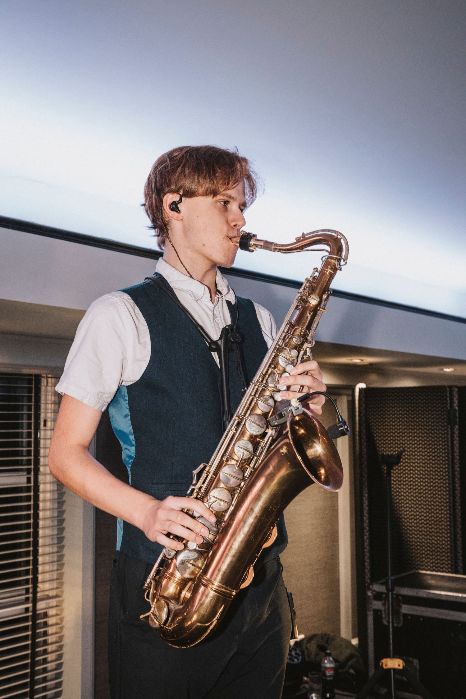 A musician plays a brass saxophone while wearing a dark vest and white shirt in a dimly lit room.