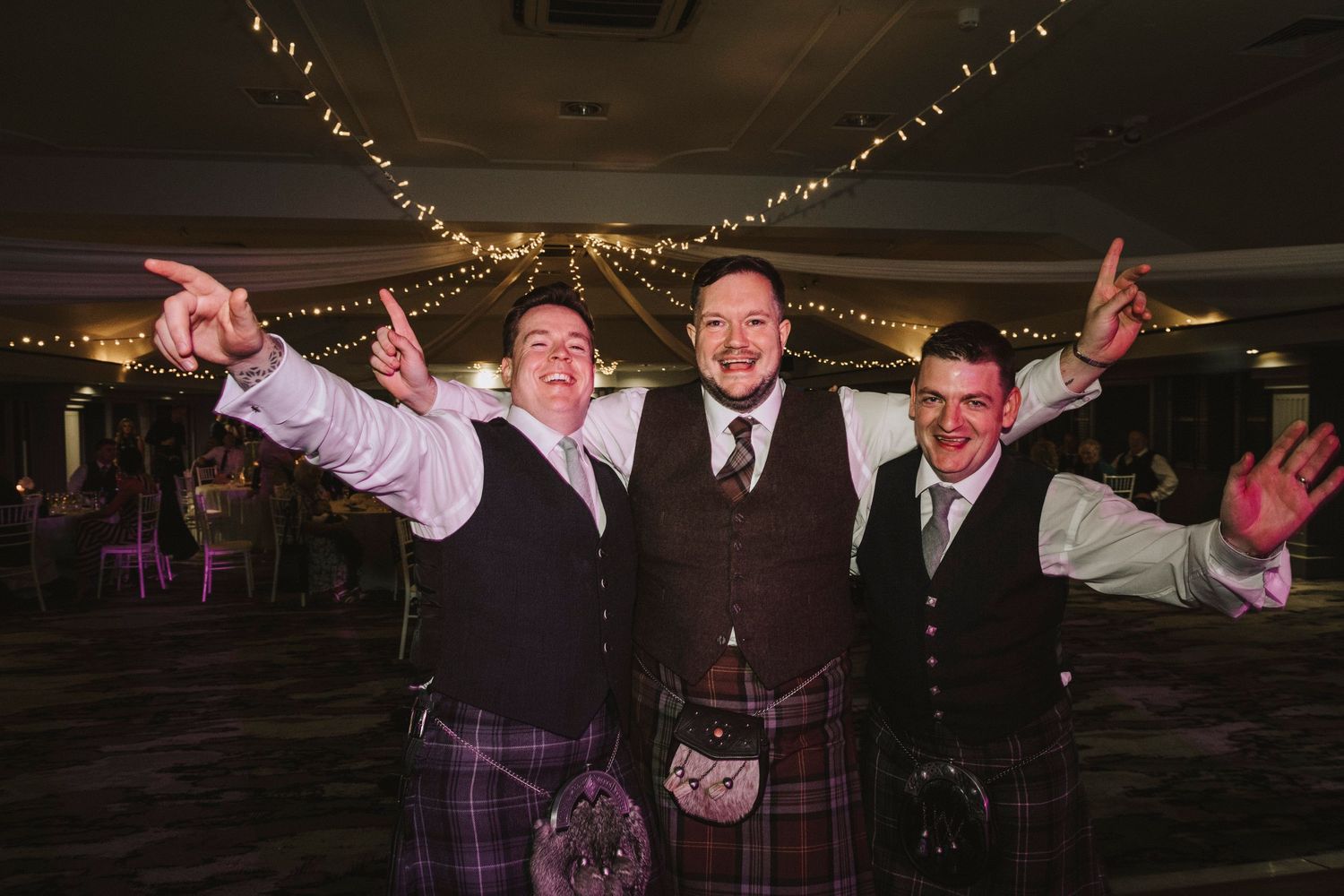Wedding party members dance and celebrate on the dance floor during an evening reception with string lights overhead.