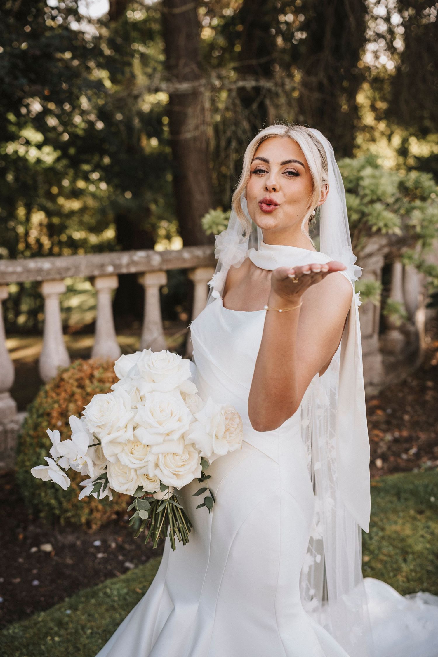 A bride in a white wedding dress and veil poses playfully in a sunlit outdoor garden setting with white roses.