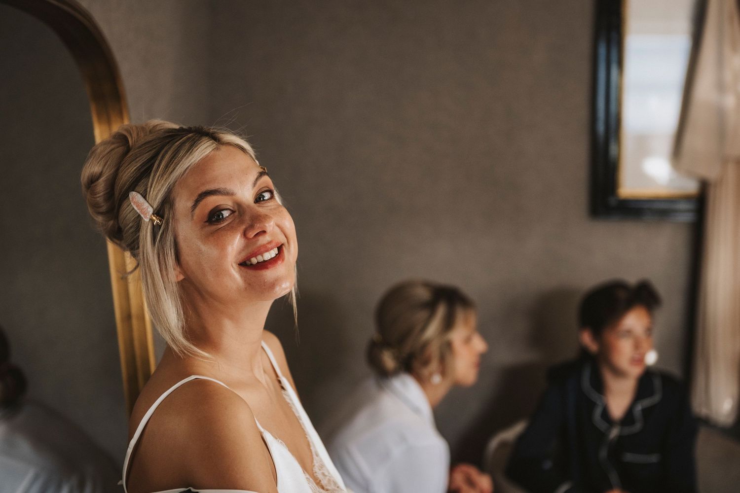 A person in a white top smiles brightly while getting ready in a room with others in the background.