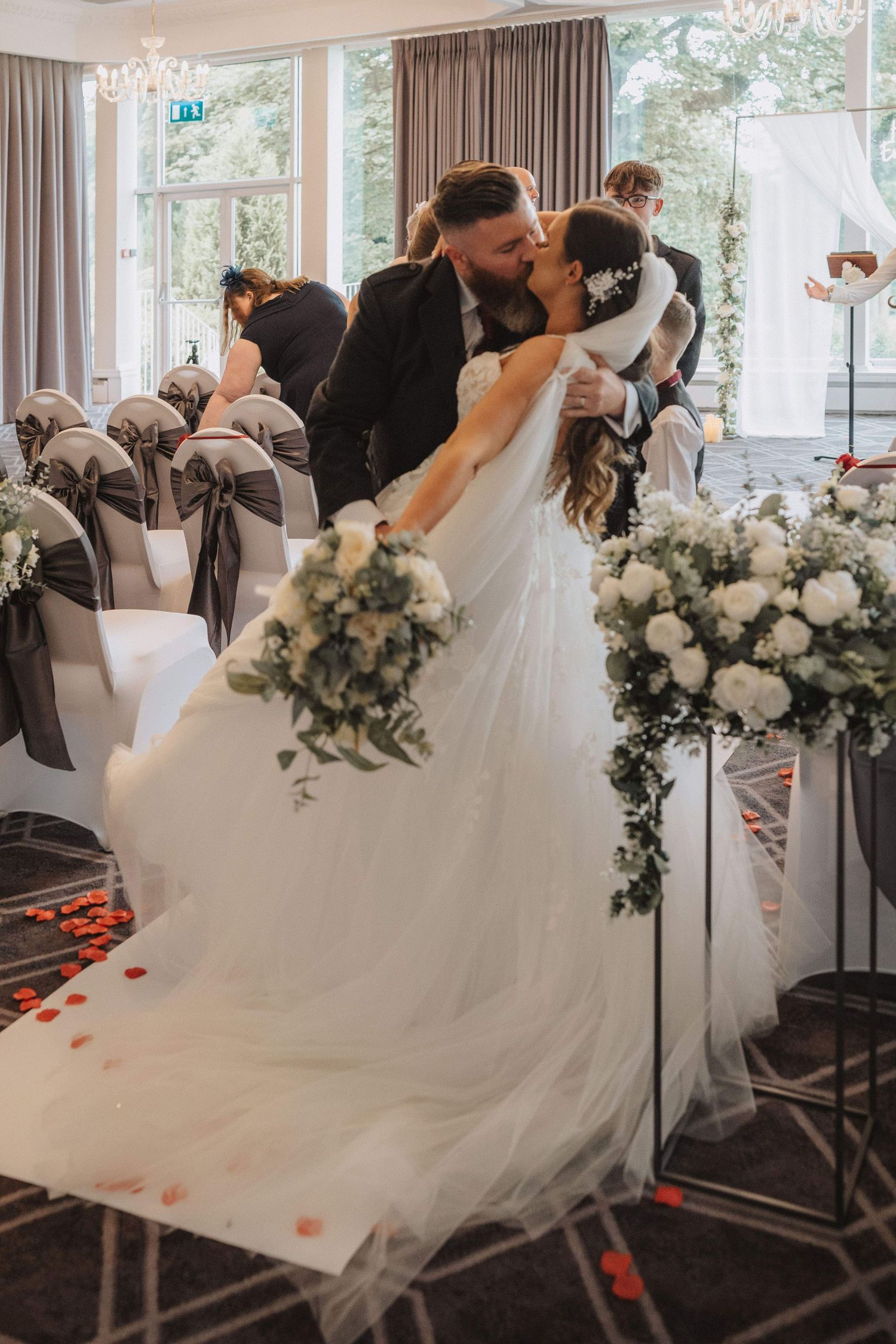 A romantic wedding kiss captured among white floral arrangements and decorated ceremony chairs.