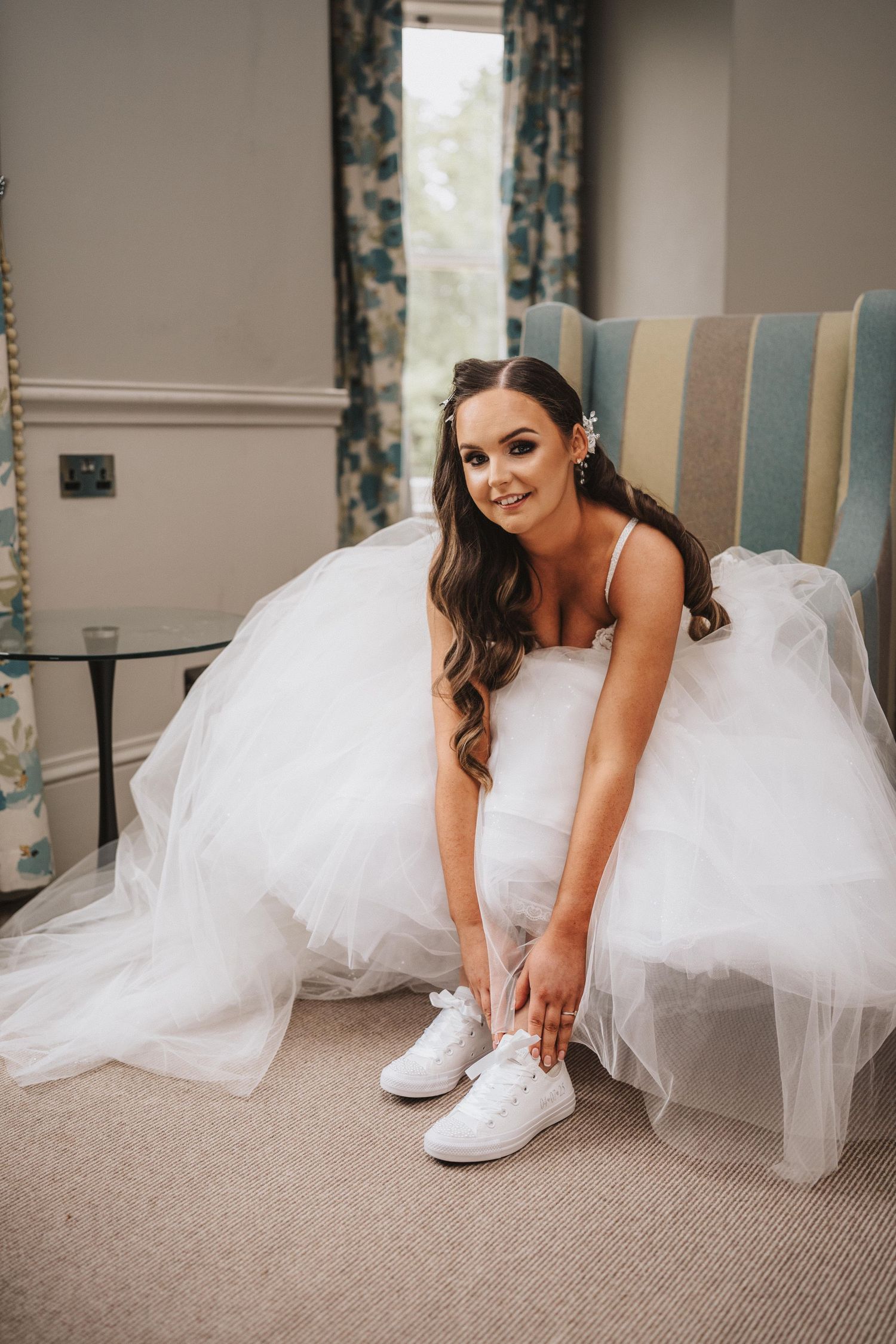 A bride in a white wedding gown sits in a chair while putting on white sneakers.