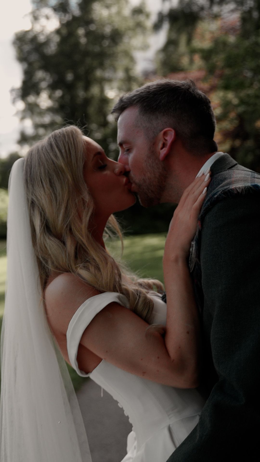 A bride and groom share a romantic kiss outdoors, with the bride wearing a white dress and veil on their wedding day.