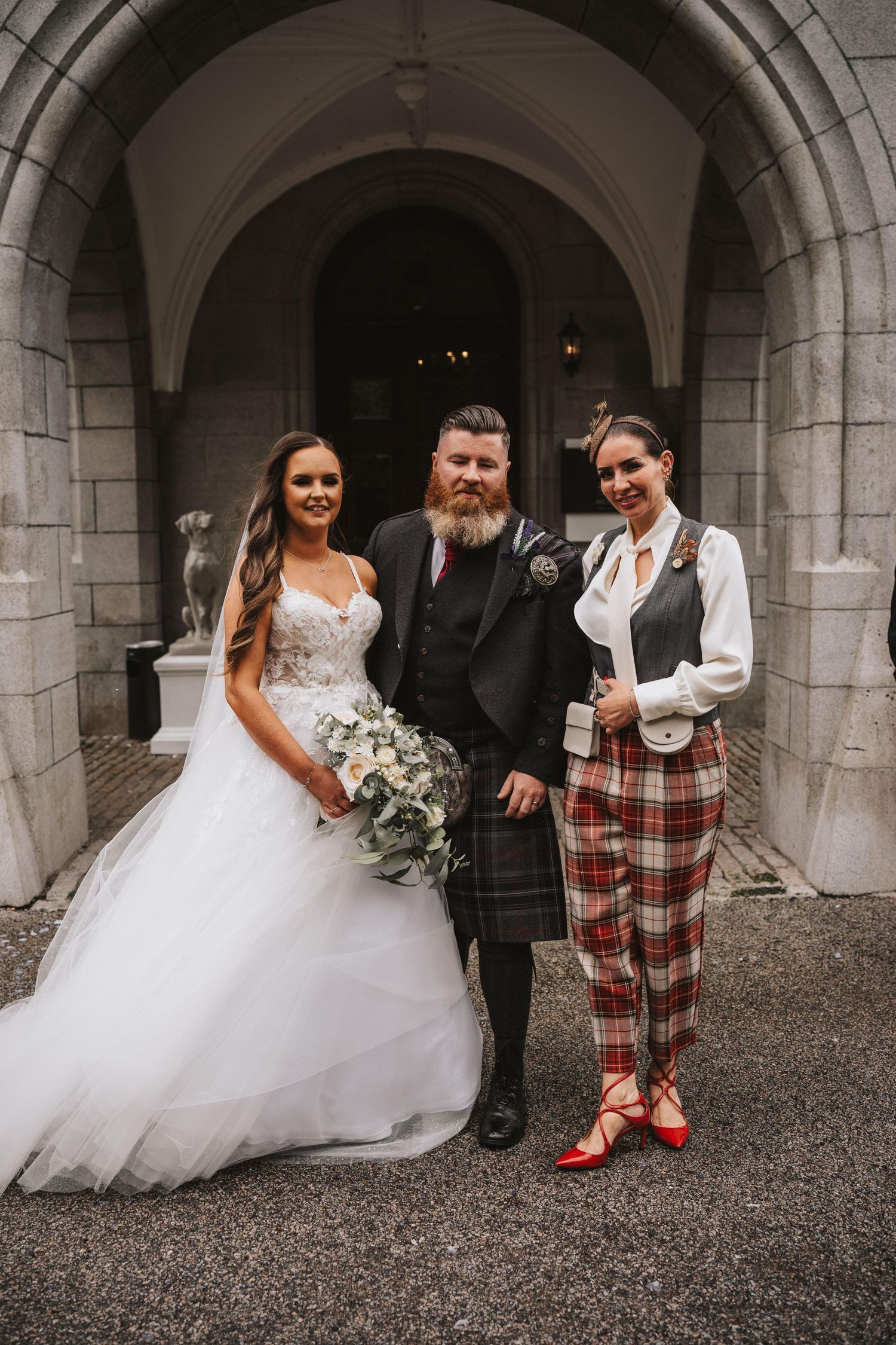 Wedding party poses under stone archways with bride in white dress and groom in Scottish formal wear.