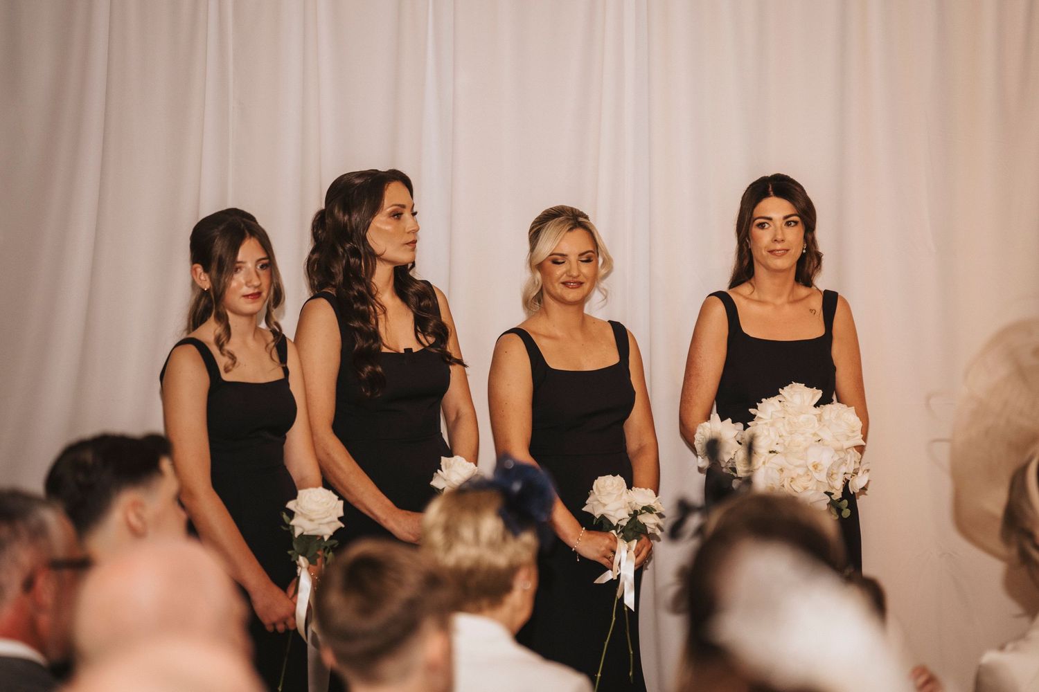 Bridesmaids in black dresses holding white bouquets stand in a line during a wedding ceremony.