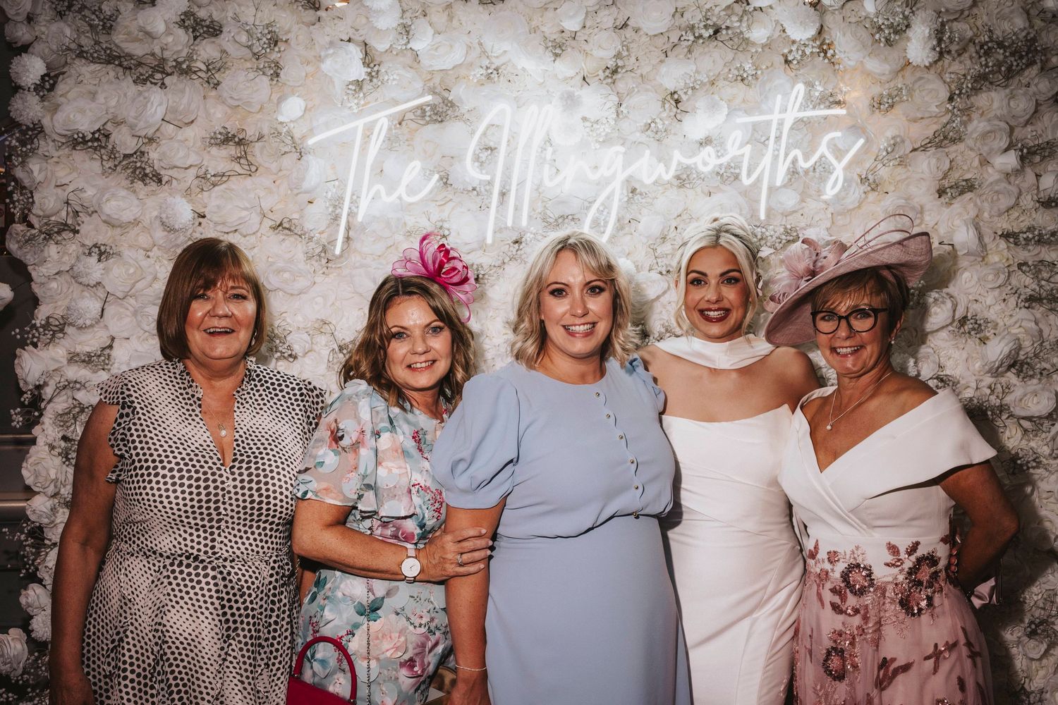 A group of women embracing and smiling together at a social gathering against a floral patterned backdrop.