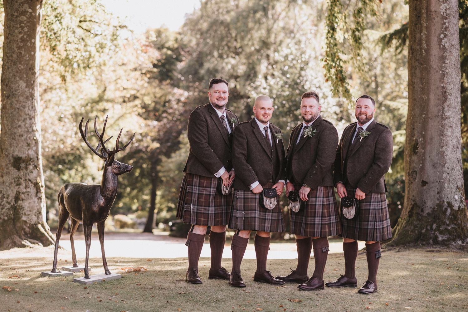 A group wearing traditional Scottish kilts poses together outdoors in a wooded setting with tall trees.