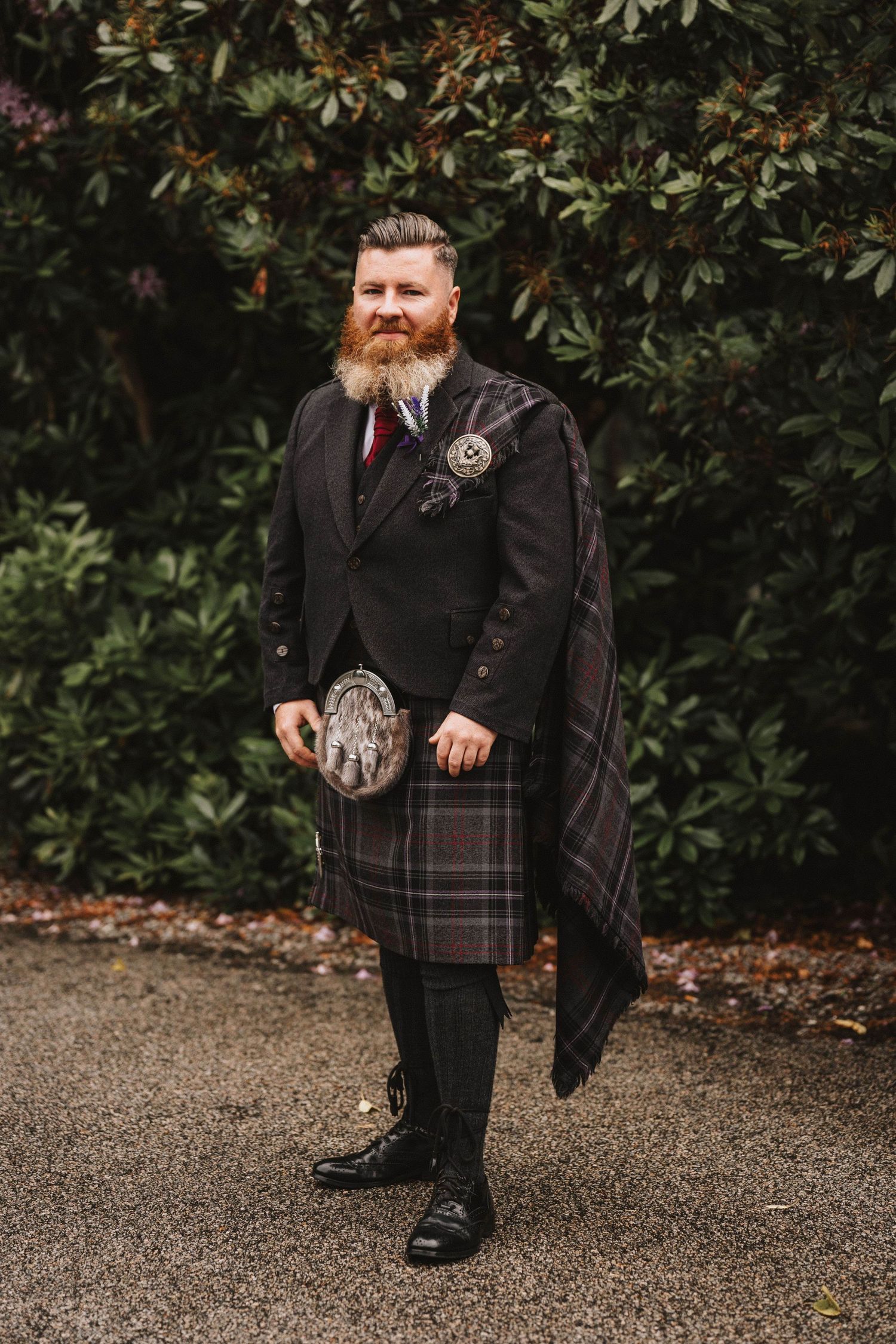 Wedding party member wearing traditional Scottish kilt and formal black jacket poses outdoors against foliage.
