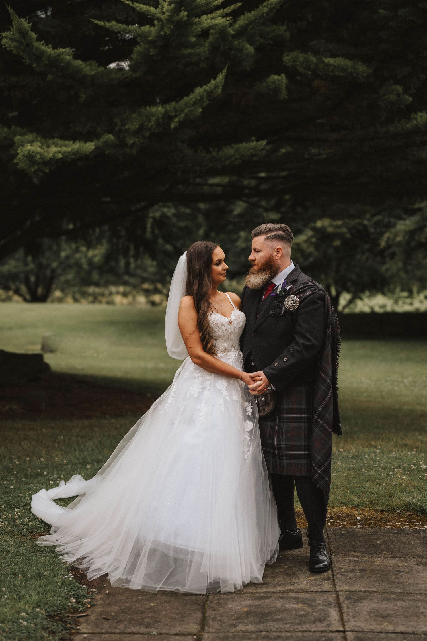 Romantic wedding portrait of couple in Scottish attire with bride in flowing white gown against evergreen trees.