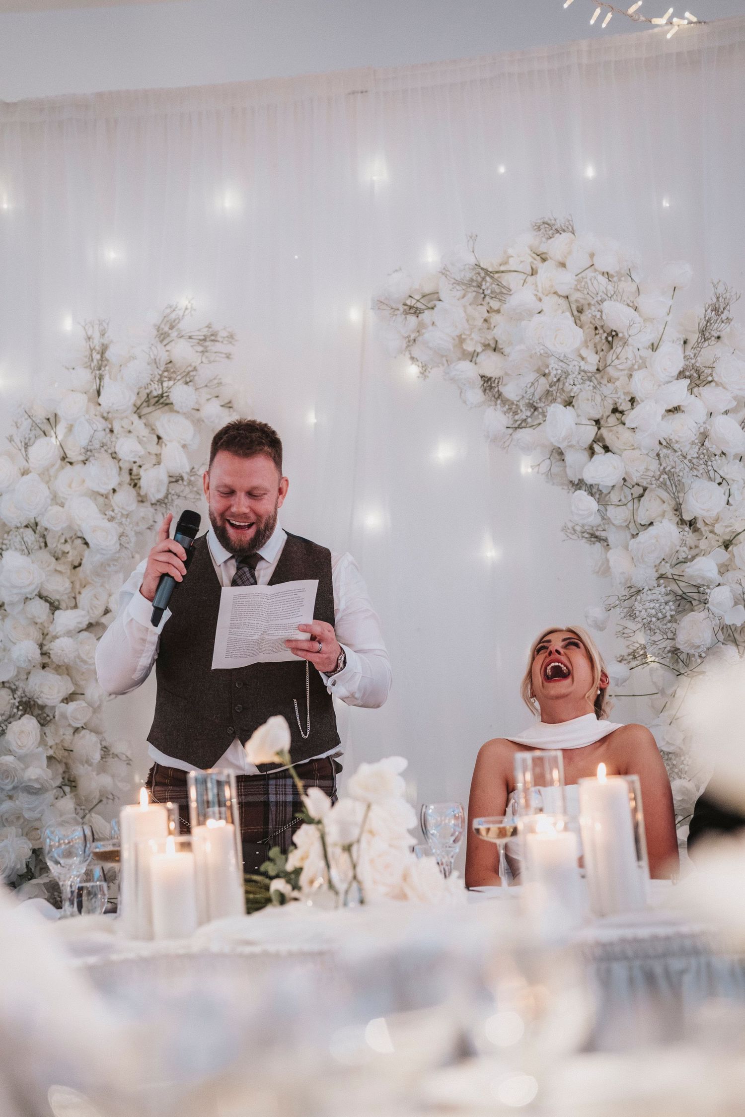 A candid sequence of joyful reactions during wedding speeches at an elegant white floral reception.