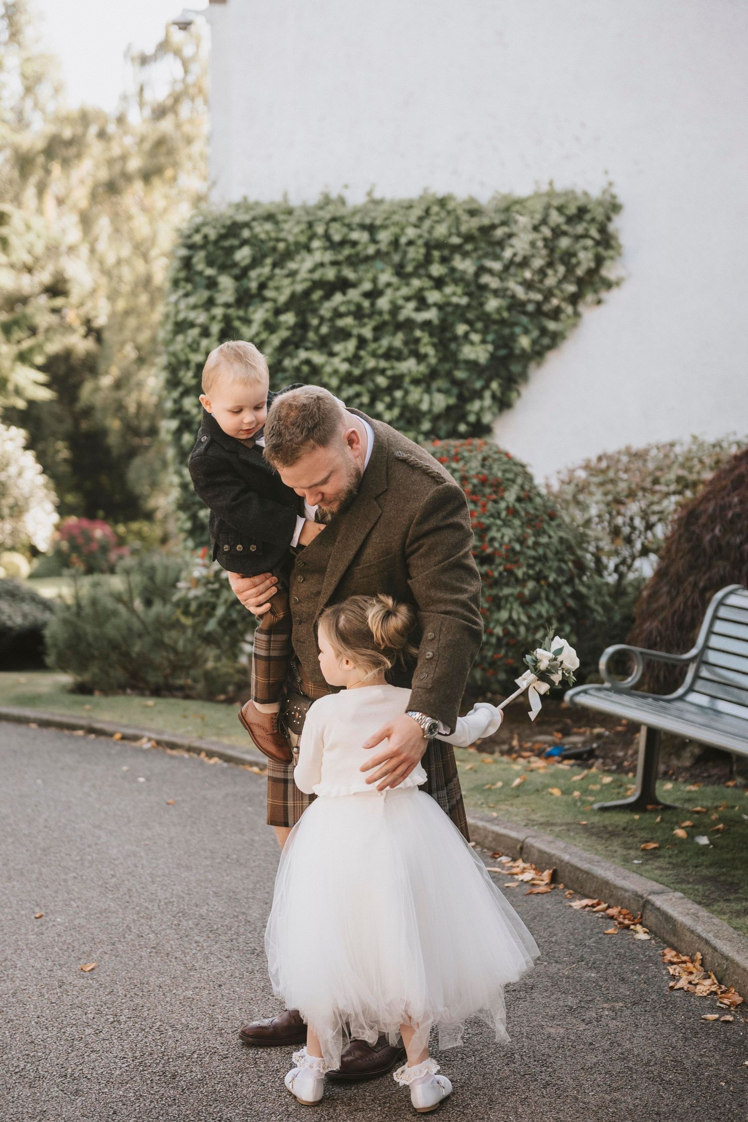 A touching moment captured in a garden setting as a figure in a brown coat embraces a small child in a white tutu dress.