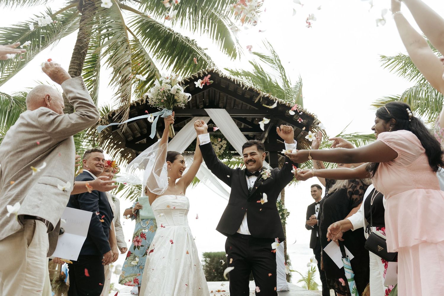 Guests cheering as newlyweds walk under floral arch – Cook Islands beach ceremony
