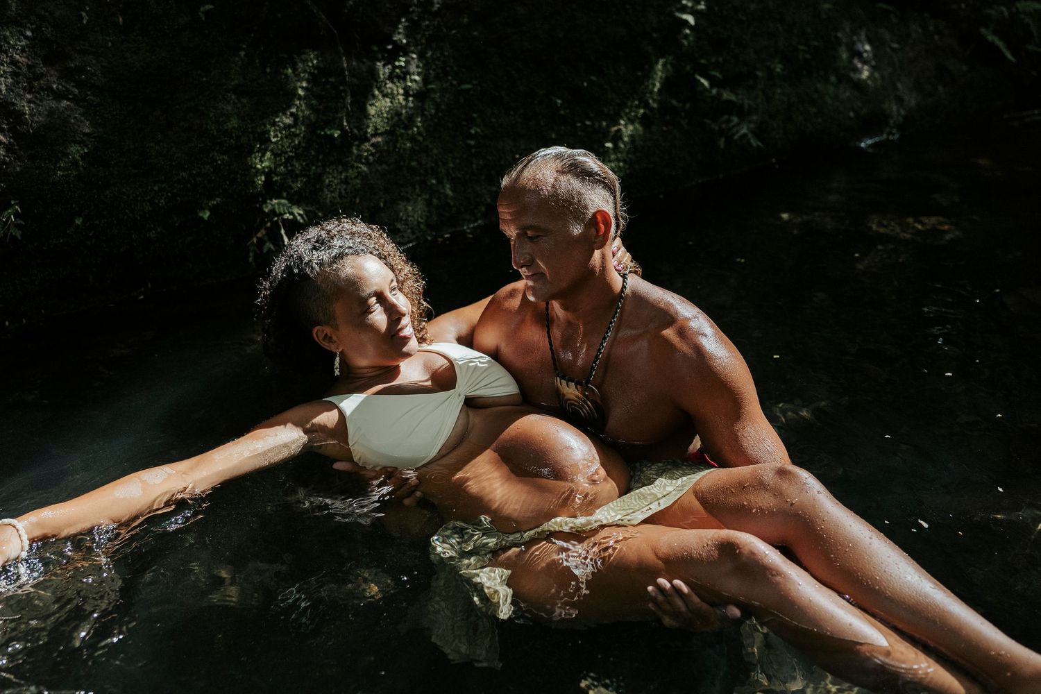 Pregnant Woman sitting on her husband in the River in Rarotonga- Moody Maternity Portrait