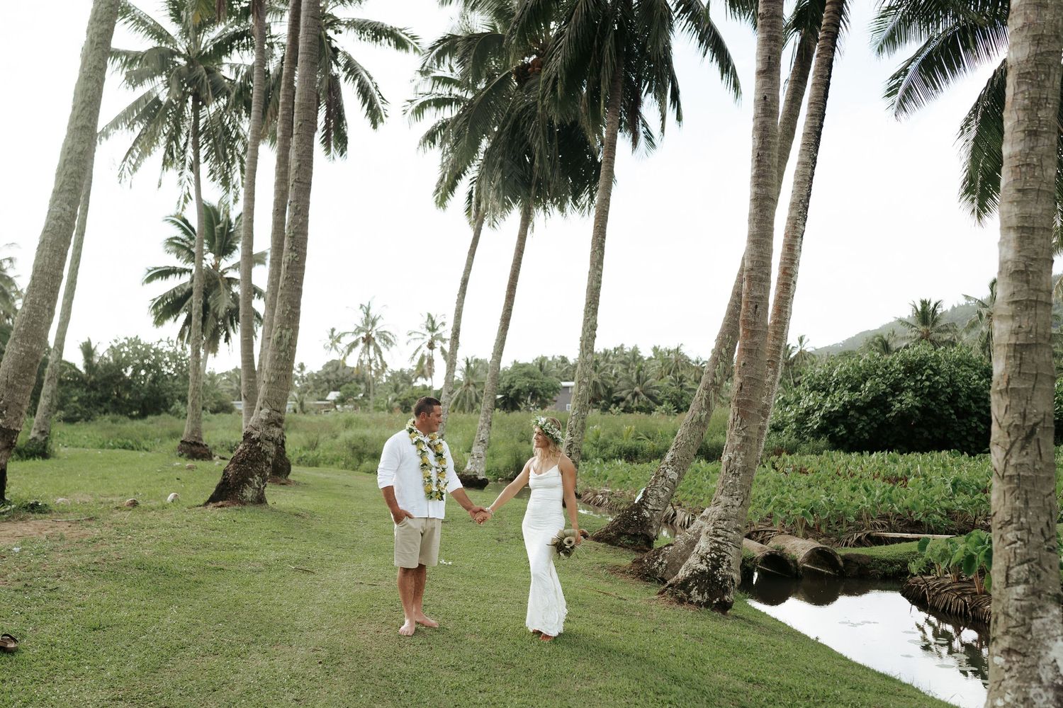 Bride and groom walking across coconut grove over tropical stream – lush island wedding location