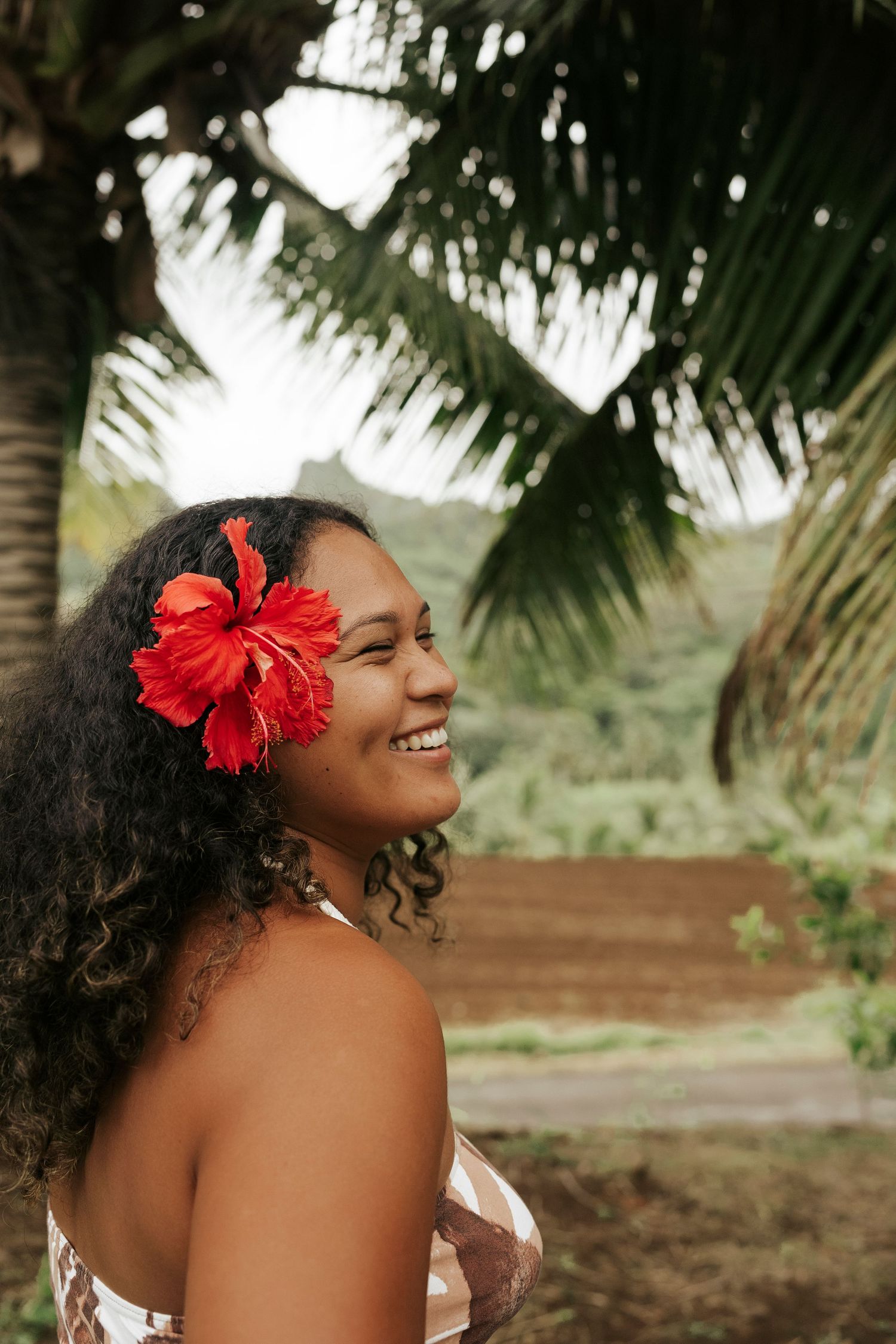 Close-up of woman with red hibiscus in her hair smiling in front of palm trees – island portrait session
