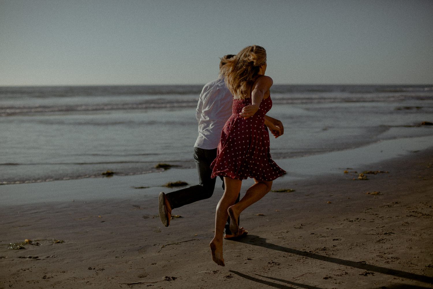 Two people playfully running along the beach at dusk with ocean waves in background.