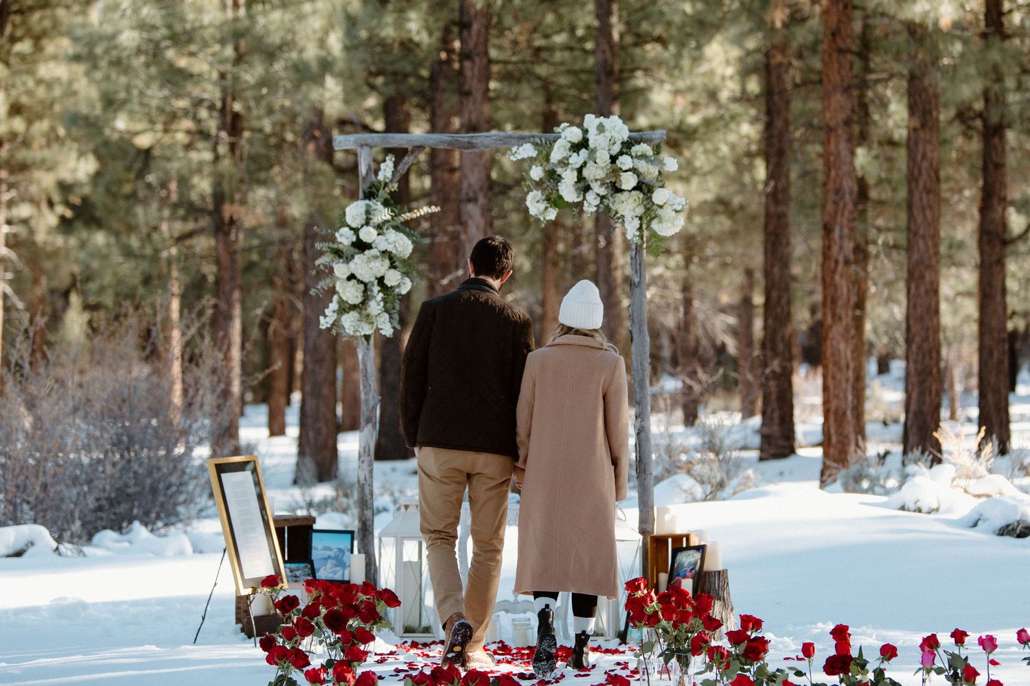 A romantic winter proposal setup with white floral arch and red roses in a snowy forest setting.