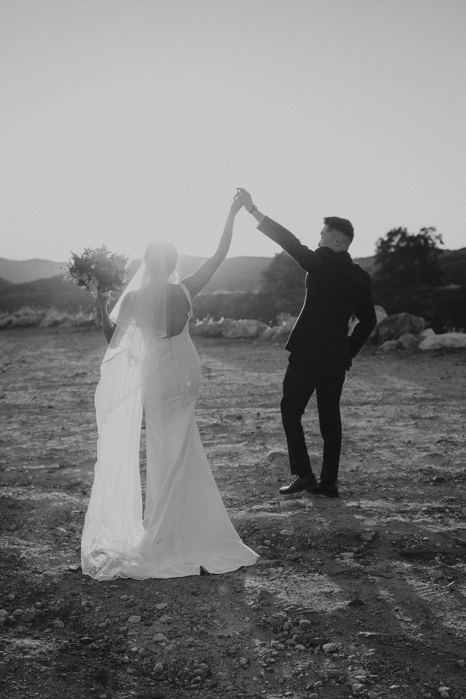 A couple in wedding attire dancing together on a hilltop at sunset.